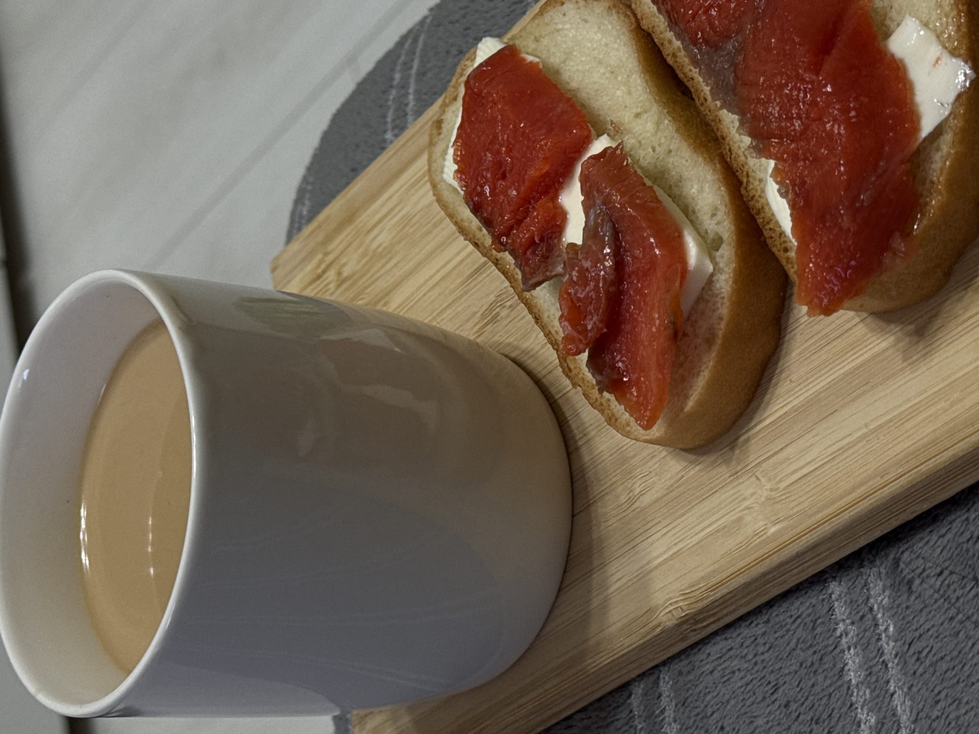 The image depicts a cup of coffee and two open-faced sandwiches on a light-colored wooden cutting board. The sandwiches appear to be on white bread with a white spread and topped with slices of red fish, possibly salmon. The cup is white and cylindrical, containing a light brown liquid. The board rests on a gray surface with subtle stripes or patterns. The location is Unknown, Unknown.