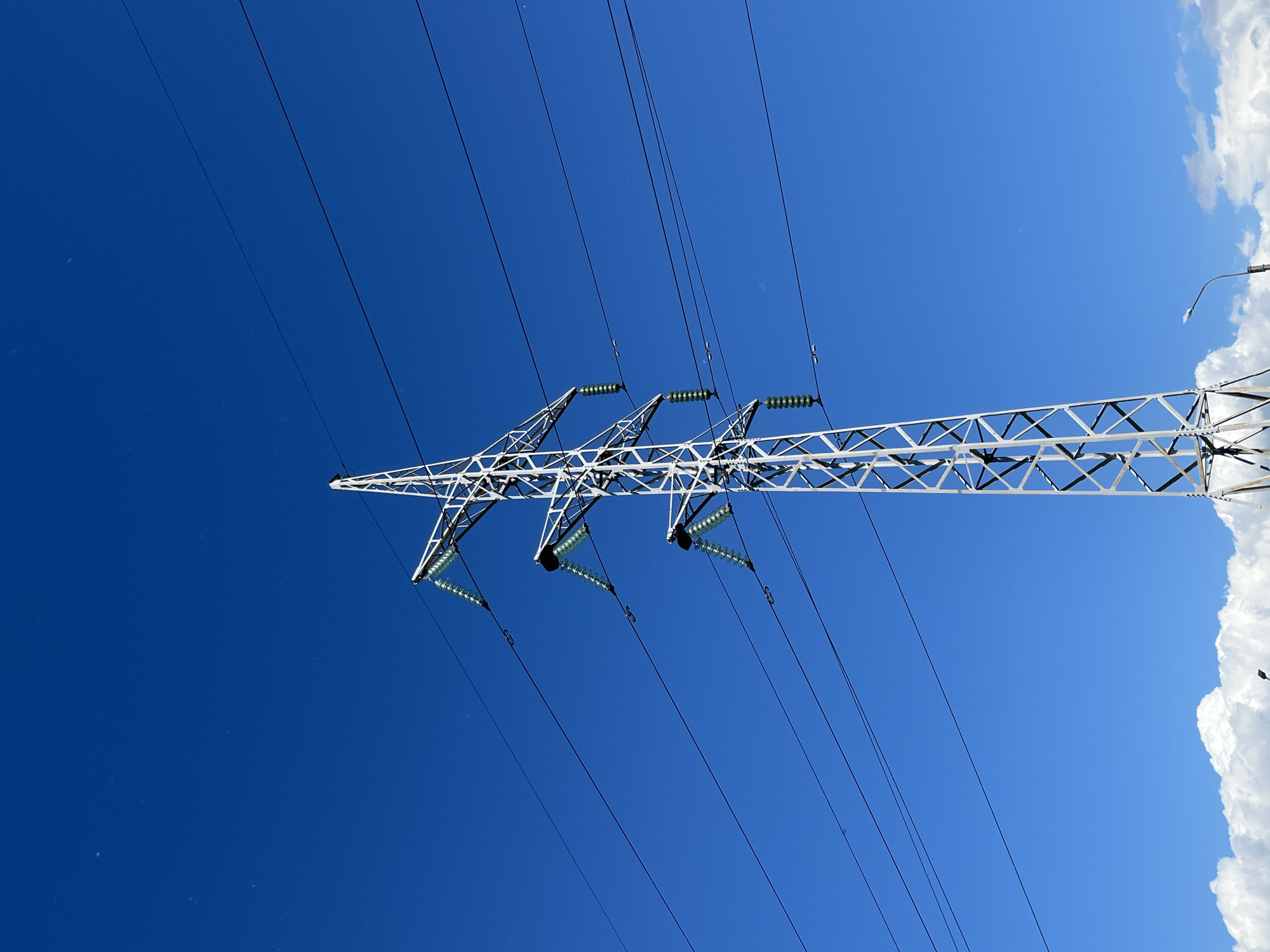 A tall, white, metal electricity pylon stands against a clear blue sky, its lattice structure visible. Several thick black power lines extend from the pylon, disappearing out of frame to the left and right. Attached to the pylon are three sets of green insulators, each holding a power line. In the lower part of the image, a line of fluffy white clouds stretches across the bottom, and a street lamp is visible on the right side. The scene is devoid of people and any discernible activity. The environment appears to be an open area, likely in or around the city of Saint Petersburg, Russia, given the presence of electricity infrastructure and a typical sky and cloud formation for the region.