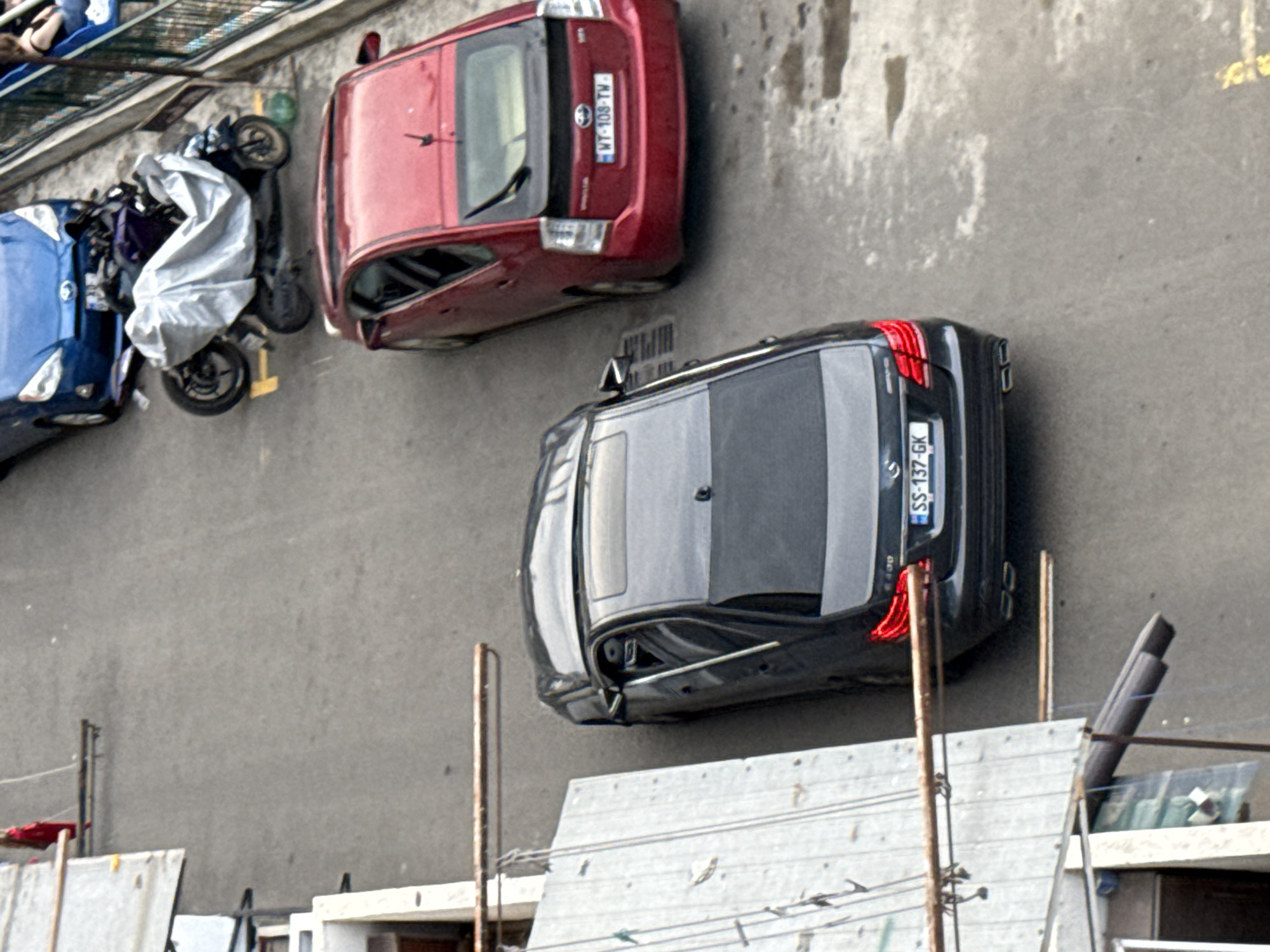 The image shows a top-down view of a street in Tbilisi, Georgia. Two cars are parked on the right side of the road. A black Mercedes-Benz sedan is parked in the foreground, with its license plate visible as "SS-137-GK". Behind it, a red Toyota Prius is parked, displaying the license plate "WT-108-TW". A motorcycle covered with a white tarp is parked to the left of the Prius, partially obscured by a blue car. In the background, a portion of a railing and a seated person are visible. The left side of the image features architectural elements including a white corrugated wall and metal structures. The ground surface appears to be asphalt with some wet patches and yellow markings.