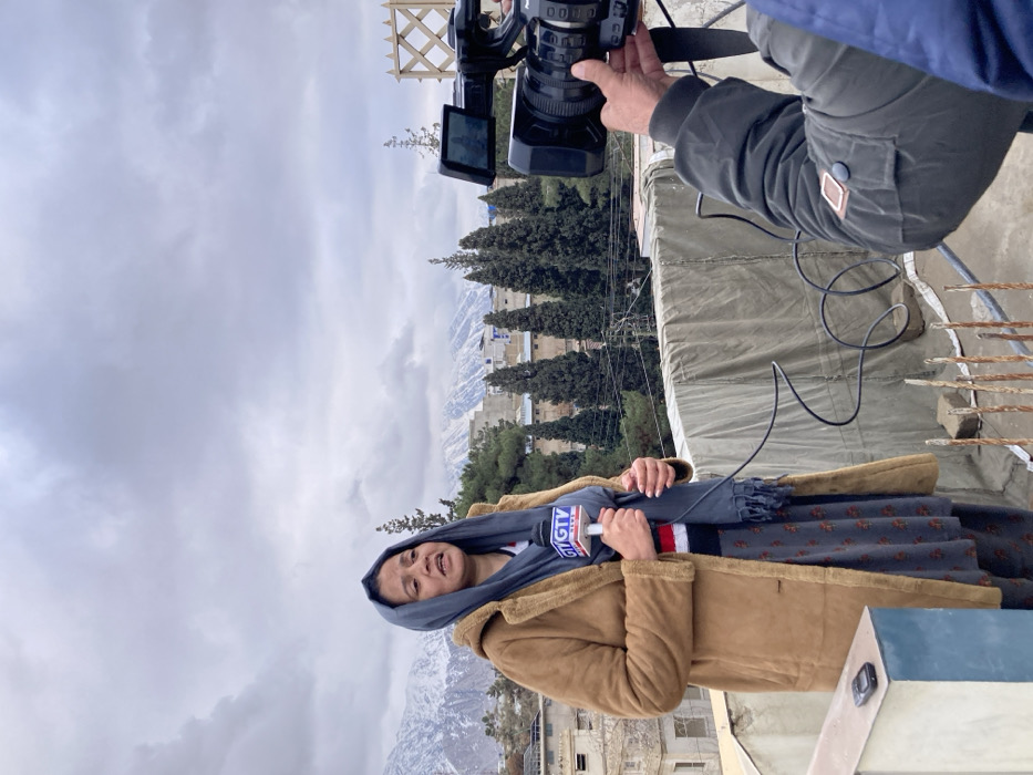 An outdoor news reporting scene is depicted on a rooftop in Quetta City Tehsil, Pakistan, under an overcast sky. In the left foreground, a female reporter stands, facing slightly right, wearing a brown long coat, a blue-grey headscarf, and a dark patterned lower garment. She holds a white microphone with a blue "GTV NEWS" logo in her left hand, appearing to be speaking. To her left, a black, older-model Nokia phone rests on a low, light-colored concrete parapet. In the right mid-ground, the right arm and hand of a male cameraman are visible, operating a black professional video camera aimed at the reporter. Black coiled cables are visible on the grey tarp-covered rooftop surface between the reporter and the camera. The mid-ground features a cityscape with multiple buildings and numerous tall, slender evergreen trees. A wooden lattice structure is partially visible on the far right. In the background, a range of snow-capped mountains stretches horizontally beneath the cloudy sky.
