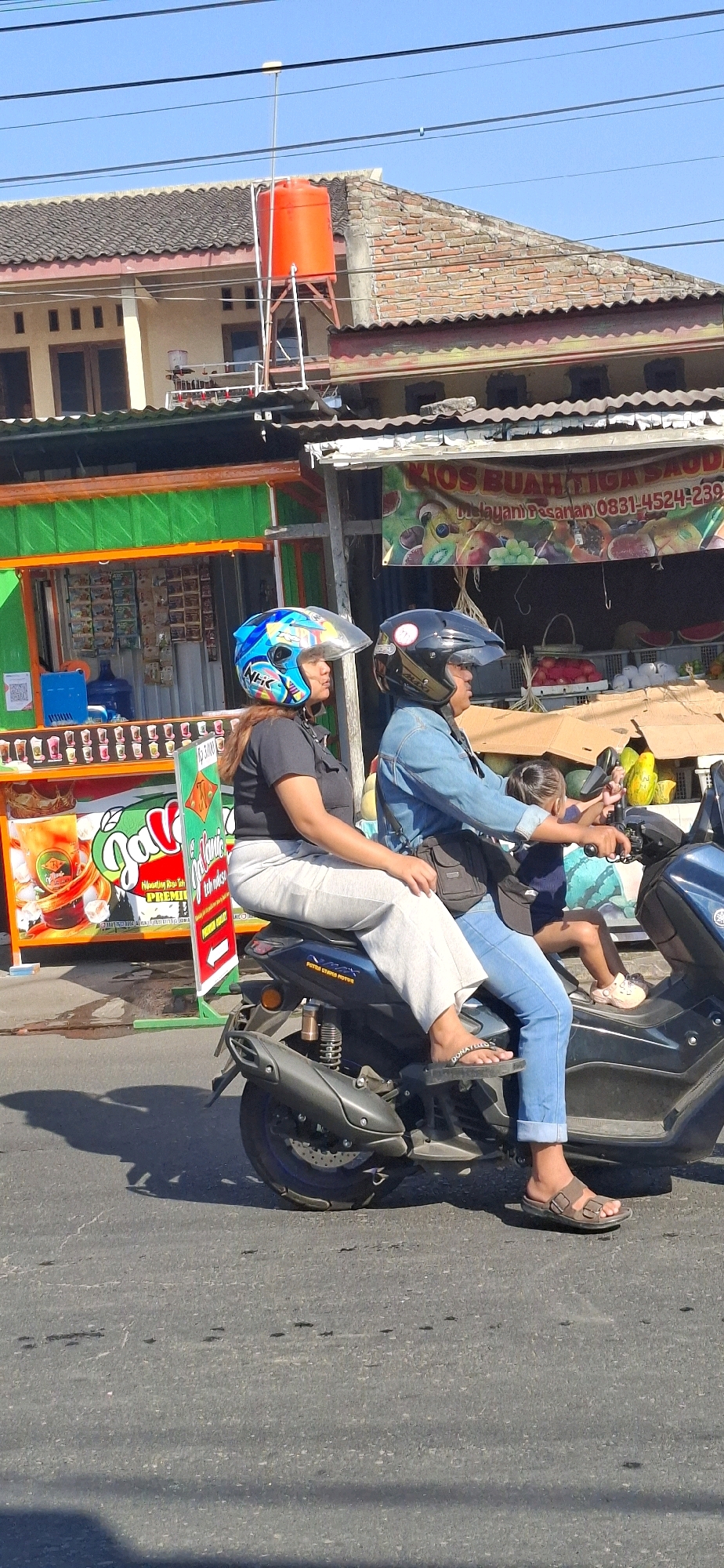 The image depicts a street scene in Desa Donohudan, Indonesia, on a clear, sunny day. In the foreground, two individuals are riding a dark-colored scooter. The rider in front is wearing a denim jacket and blue jeans, with a black helmet. The passenger behind, a woman, is wearing a black shirt and light-colored pants, with a blue and white helmet. A young child is seated between them on the scooter. Behind the scooter, there are two roadside stalls. The stall on the left is brightly colored with green and orange accents, displaying various products on shelves. The stall on the right has a banner with text and images of fruits, and it appears to be a fruit stand. The background shows buildings with tiled roofs and a bright blue sky. An orange water tank is visible on top of one of the buildings. The road surface is asphalt, with some cracks and debris. The overall impression is a snapshot of daily life on a street in a populated area of Indonesia.