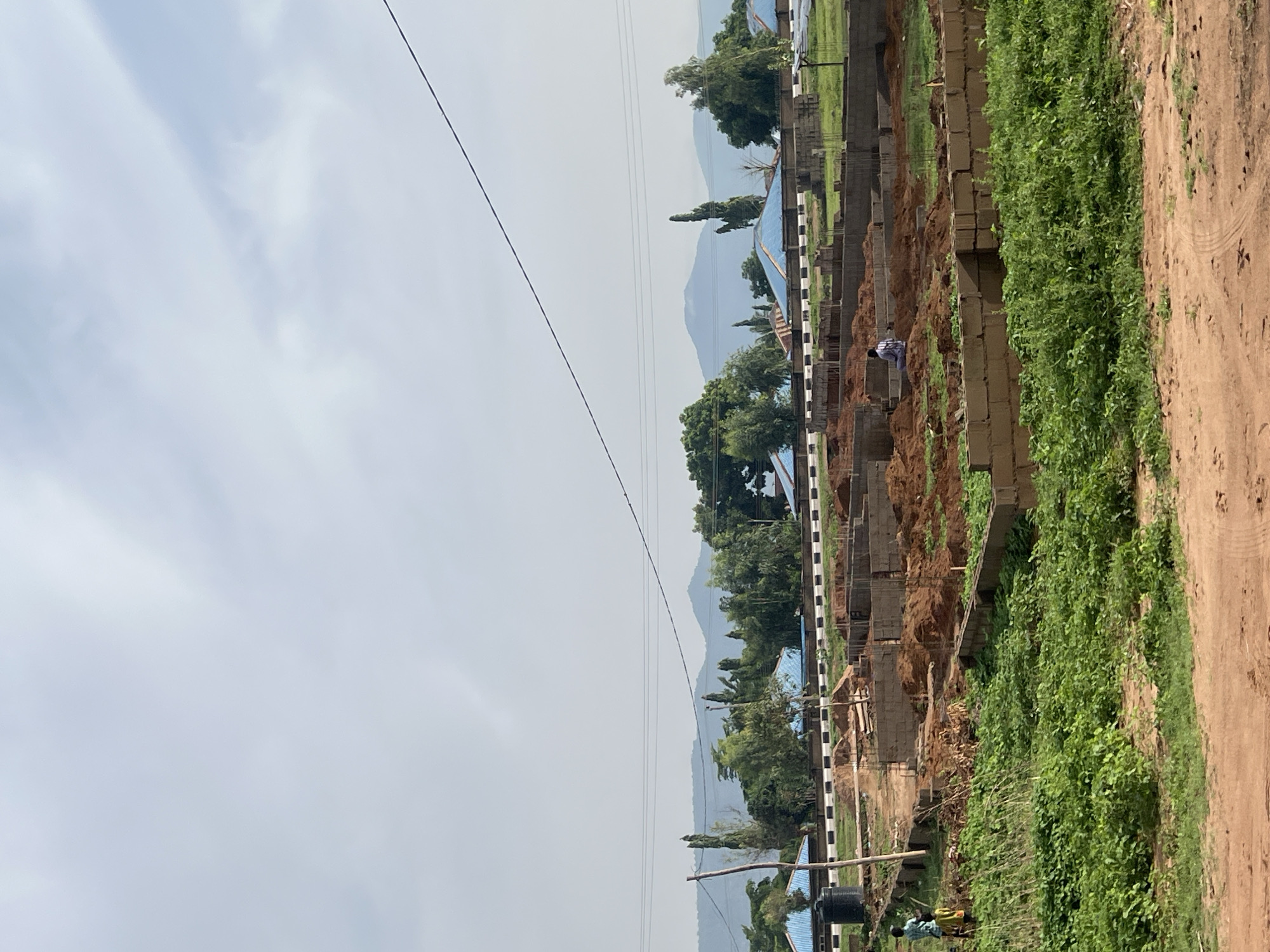 The image shows a construction site in Howayi, Nigeria. There are several unfinished buildings with visible brickwork and soil piled up around them. A dirt road runs along the edge of the site, adjacent to lush green vegetation. In the background, mountains are visible under a hazy sky. Power lines run across the scene. Two individuals stand near a water tank at the lower-left corner of the image. A person in a blue and white patterned shirt is present within the construction area.