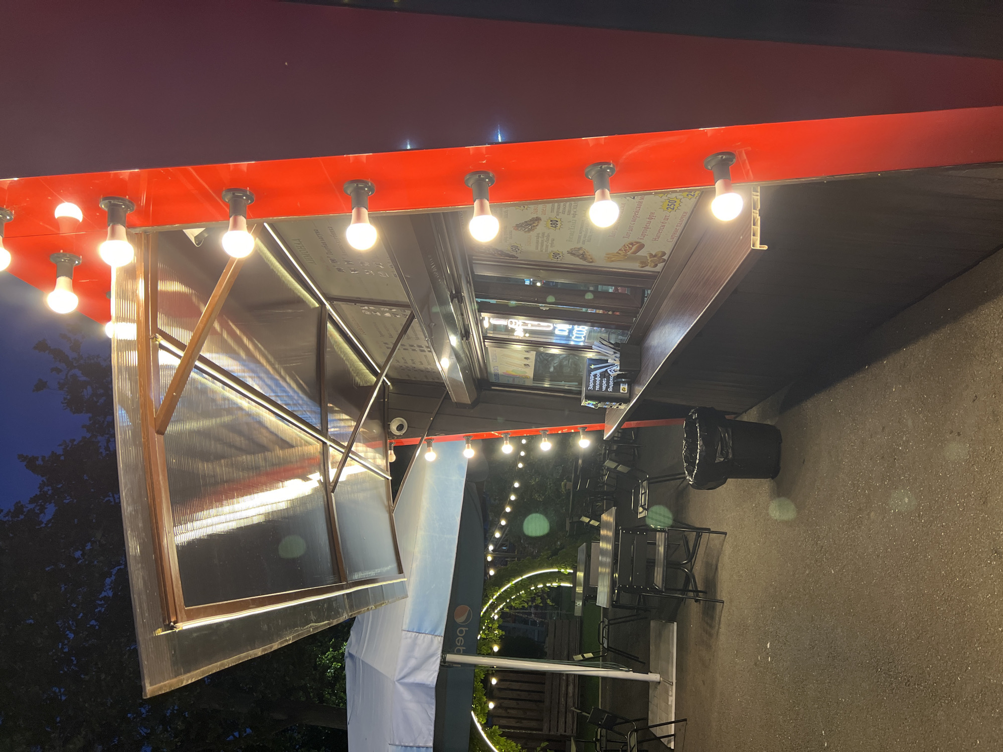 The image shows an outdoor food stall at night, likely in the city of Krasnodar, Russia. The stall is red with multiple light bulbs lining its side. It has a clear awning covering a service window. Inside the window, menus are visible, displaying food items like hot dogs and french fries. To the left of the stall, there are tables and chairs set up for outdoor dining, along with a large umbrella and decorative string lights in a semi-circular arch shape. The ground is paved with asphalt, and a trash can is located near the stall. Trees and foliage are visible in the background, and the sky is dark, indicating nighttime. No people are present in the image.