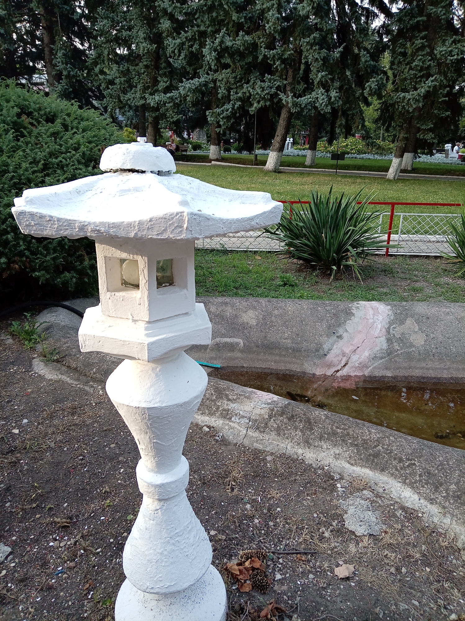 The image depicts a white, pagoda-style garden lantern situated in the foreground. The lantern is made of concrete or a similar material and has a tiered roof and a square body with glass panes. Behind the lantern, a concrete channel containing murky water is visible. The surroundings include gravelly ground, green bushes, and tall evergreen trees. In the background, there is a patch of grass, a red and white fence, and more trees. The scene appears to be in a park or garden setting. The provided context indicates this location is in the city of Yessentuki, Russia. No people are prominently featured or actively engaged in any discernable actions. The overall impression is of a tranquil outdoor space, likely a park.