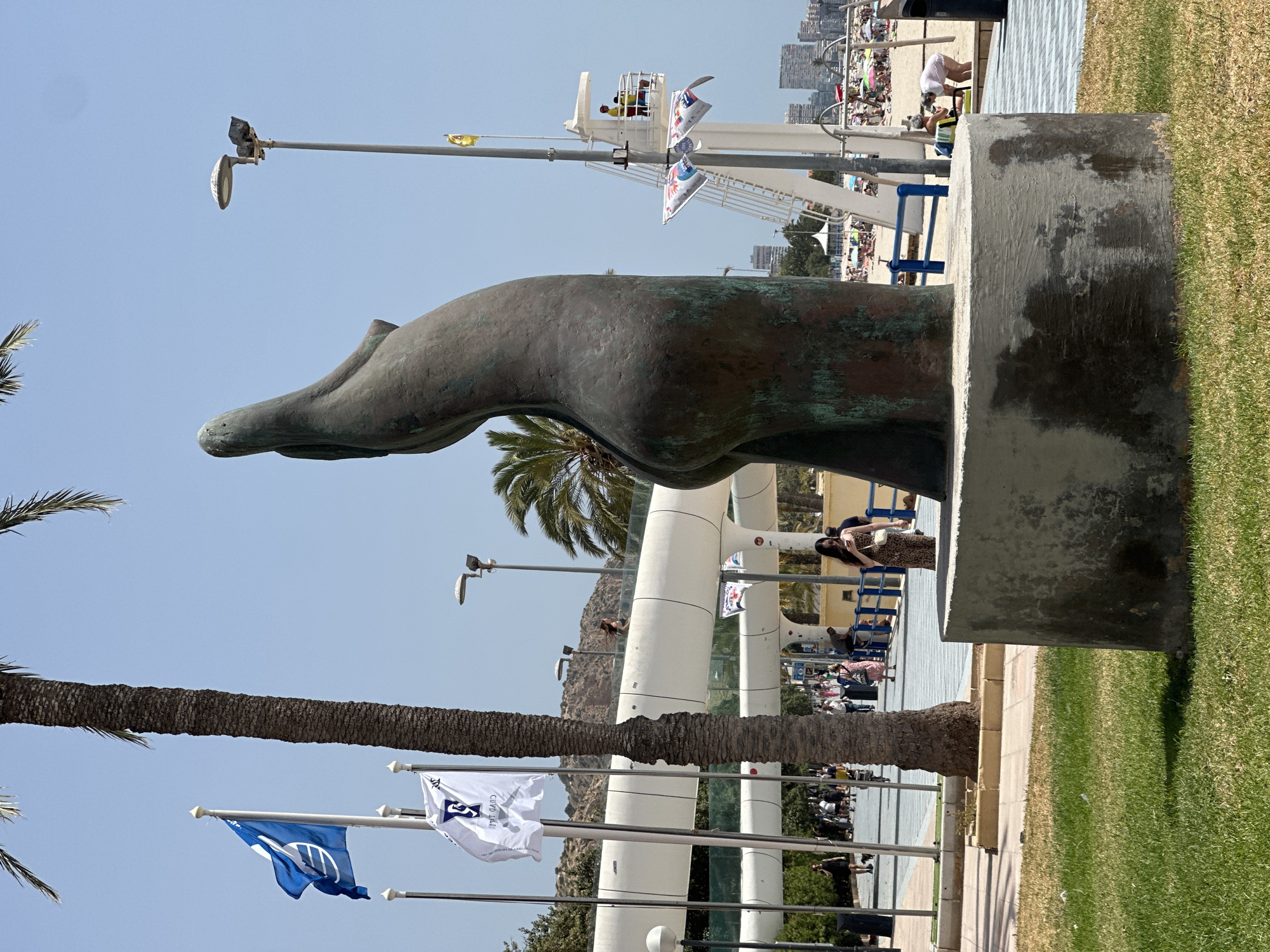 A bronze statue of a woman's torso, tilted upwards, stands on a concrete base in a grassy area. The statue is weathered and green in places. Behind the statue, a palm tree trunk is visible. In the background, a modern, curved, white walkway spans across the scene. Several flags are displayed on poles, including a blue flag with a white circle and a white flag with a blue emblem. Further back, a beach with numerous people, umbrellas, and buildings in the distance can be seen under a clear blue sky. A lifeguard tower with two people in yellow vests is also visible on the beach. People are walking along the walkway and on the beach.