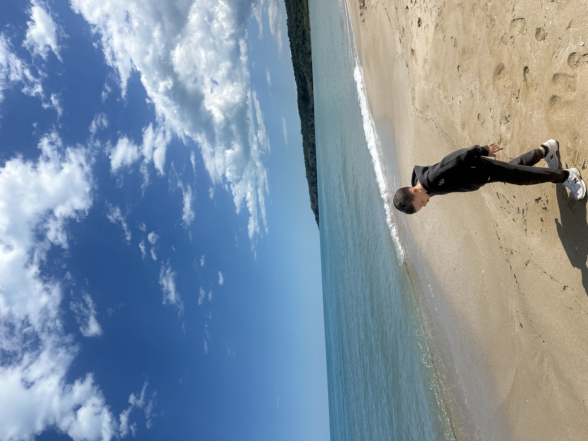 The image shows a beach scene, likely in Sozopol, Bulgaria, given the vegetation. A young person stands on the sand near the water's edge, wearing a black hoodie and pants, and light-colored sneakers. Their shadow is cast on the sand. The beach is light brown with visible footprints. The water is blue-green with small waves lapping the shore. Beyond the water is a strip of green trees and vegetation. The sky is bright blue with scattered white clouds.