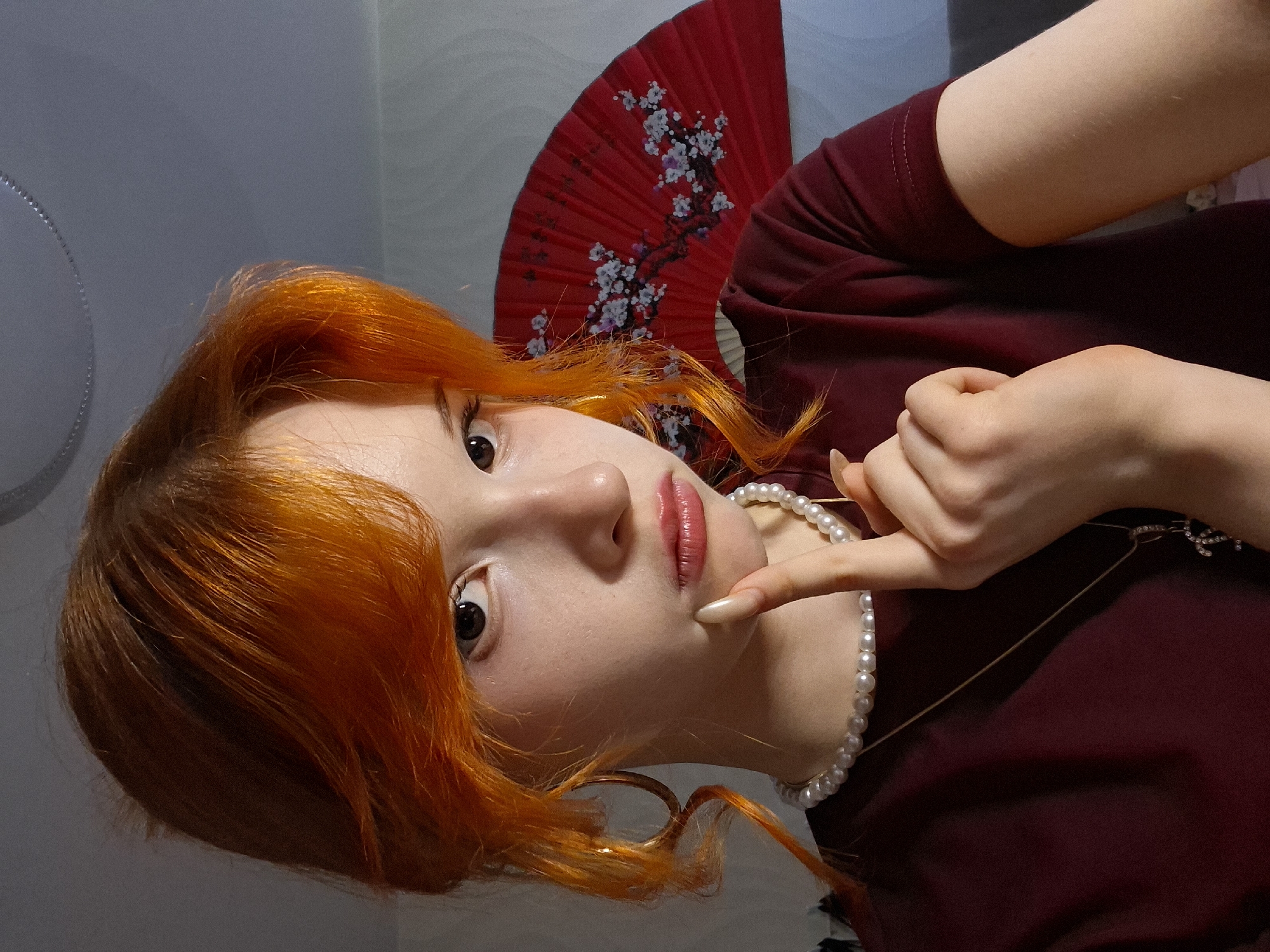 A young woman with bright orange hair is posing for a photograph. She is wearing a maroon-colored shirt and a pearl necklace. Her finger is touching her chin. Behind her is a red fan with white flower designs. The background is a light gray or white wall. The scene appears to be indoors. There is no specific information in the image to suggest the location is Chaikovsky, Russia.