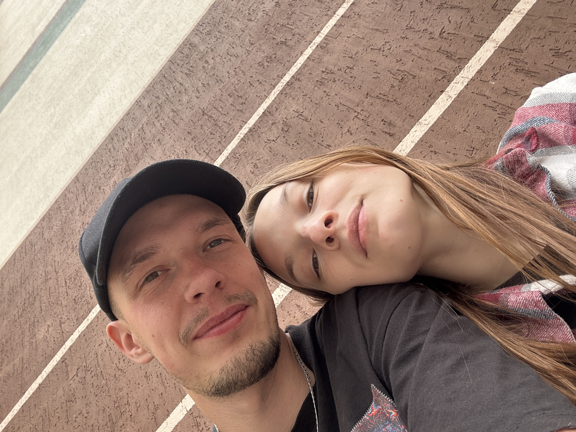 In a close-up selfie, a young man and woman are pictured from the chest up. The man, wearing a black baseball cap and a black t-shirt, is on the left, with his head tilted and a slight smile. He has a light beard and is wearing a silver chain. The woman, on the right, is leaning her head on his shoulder. She has long, light brown hair and is wearing a plaid shirt with red, white, and grey stripes. They are positioned against a textured, brown wall with diagonal white stripes. The overall setting appears to be outdoors, likely in an urban environment, possibly in the city of Chaikovsky, Russia, given the specific backdrop.