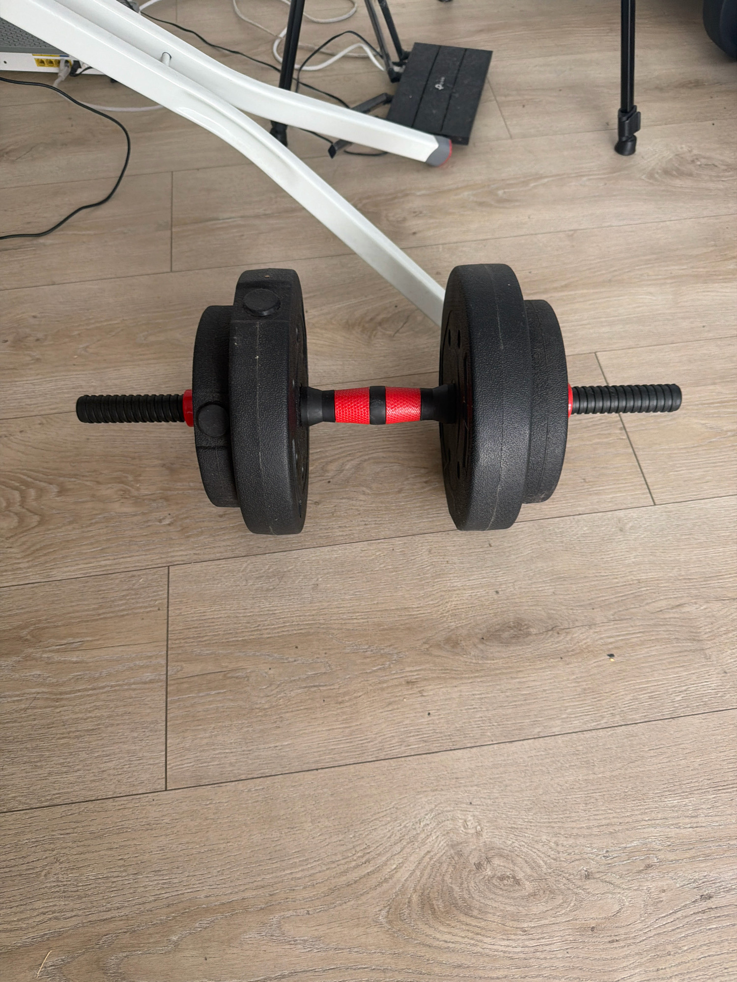 A dumbbell lies horizontally on a light brown wooden floor. The dumbbell has black weight plates on either end of a black bar with a red grip in the center. The weight plates appear to be made of plastic or coated metal. There are cables and a white stand visible in the background, suggesting an indoor setting. The scene is indoors, likely in a home or gym. The specific location within Wrocław, Poland cannot be determined from the image alone.