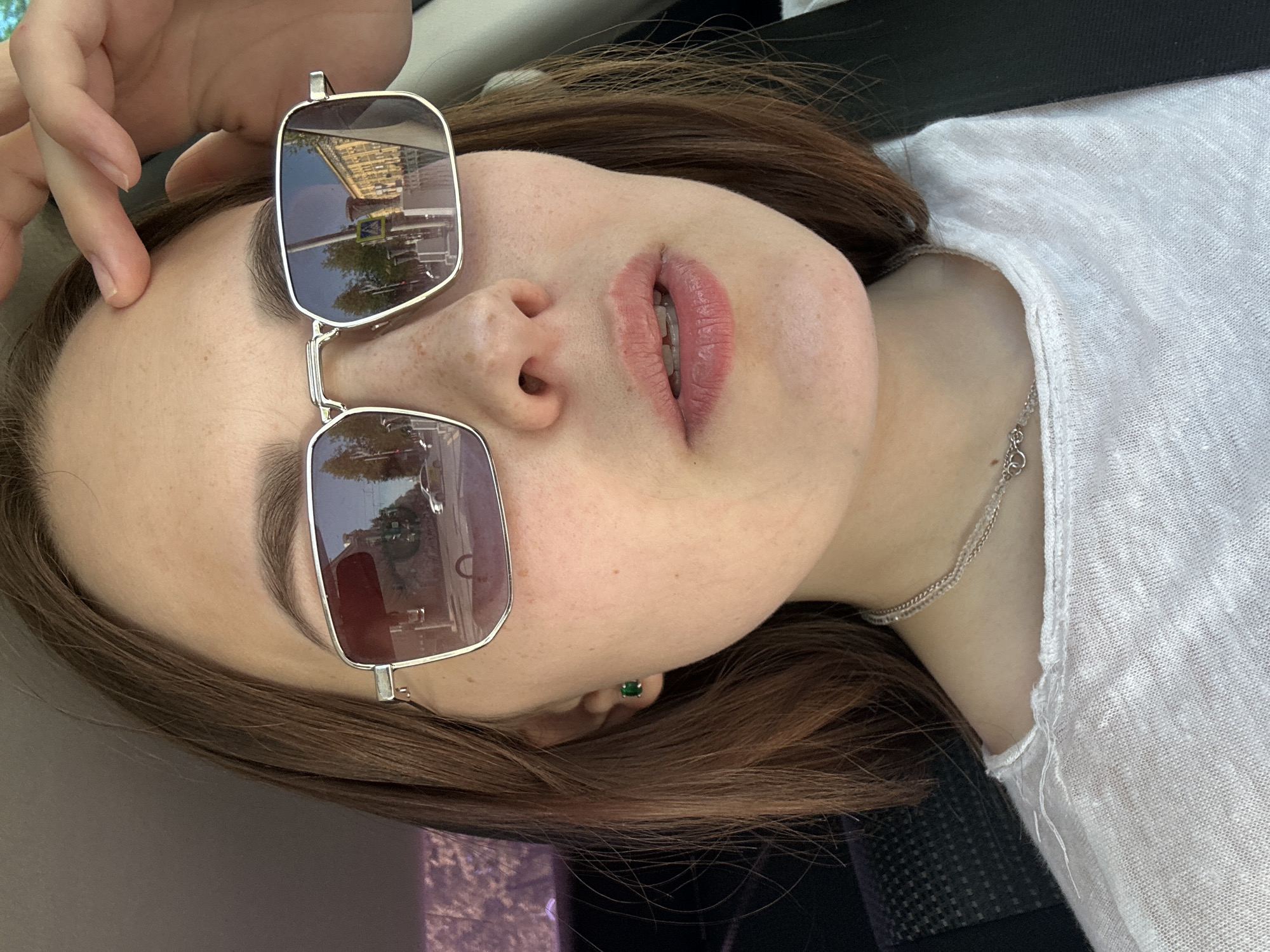A young adult female with fair skin and light brown hair is angled toward the camera, appearing to lie down. She is wearing sunglasses propped on her forehead with a silver frame, and the lenses reflect buildings and trees, indicating an outdoor location. She is wearing a white, light-colored shirt, and a silver necklace is visible on her chest. In her left ear, a small, round green earring is visible. A hand is partially visible in the upper left corner of the frame, with fingers touching her forehead. The background is out of focus but suggests a car interior.
The reflection in the sunglasses show a yellow building with multiple windows, trees and other vehicles. Using image search, the building is located at Комсомольский проспект, 79, Пермь, Пермский край, Russia, 614045.