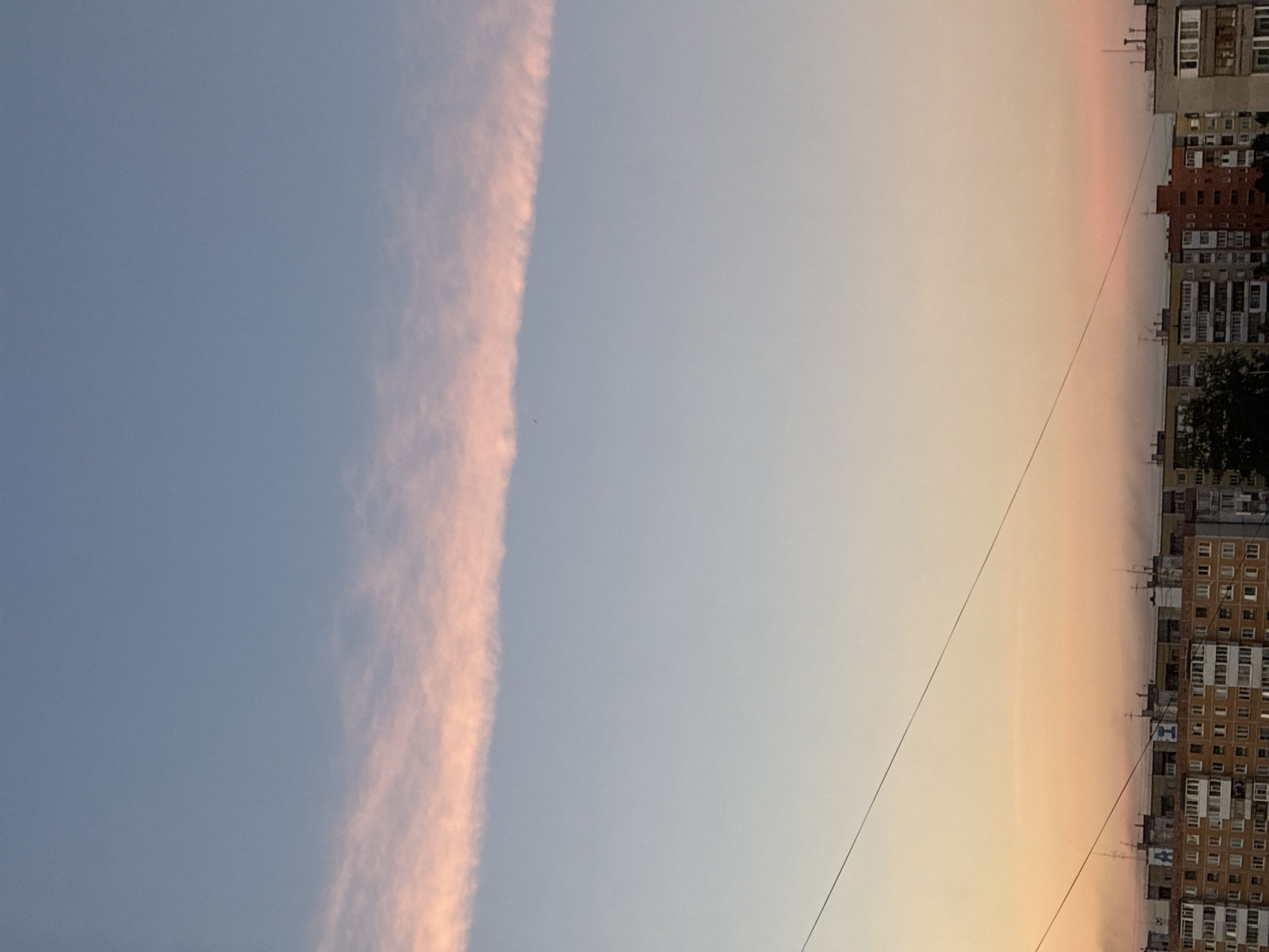 A horizontal streak of pale pink clouds stretches across a pale blue sky. Below the clouds, the sky transitions to a gradient of soft orange and yellow, hinting at a sunset. A thin black wire stretches diagonally across the frame from the bottom left to the top right, interrupting the sky. Further down, the upper portions of apartment buildings are visible, their facades a mix of brick and grey concrete. Some buildings have balconies and many windows. Several antennas and satellite dishes protrude from the rooftops. A few trees are visible to the left of the buildings. The scene appears to be an urban landscape at dusk.