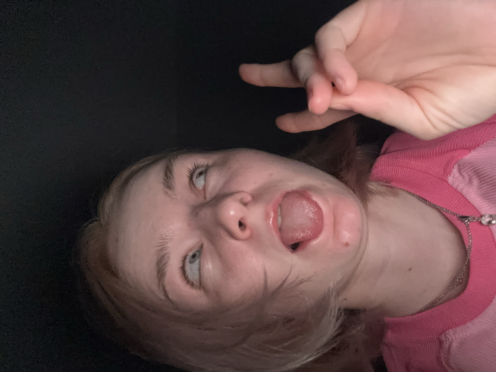 A rotated, close-up portrait features a young individual, likely female, with light skin, light eyebrows, and light-colored hair. The person is captured mid-expression, making a playful or humorous face with their eyes rolled upwards, revealing the whites, and their tongue playfully sticking out. Their left hand is visible in the upper right of the frame, forming a "peace sign" gesture. The individual is dressed in a pink garment, possibly a sweater or t-shirt, with a visible ribbed texture around the neckline. A silver-colored chain necklace with a small charm is worn around their neck. The background is uniformly dark, indicating an indoor setting with low light or a dark backdrop. The overall impression is one of casualness and lightheartedness. There is no visible text in the image. The location context provided is Vsevolozhsk, Russia.