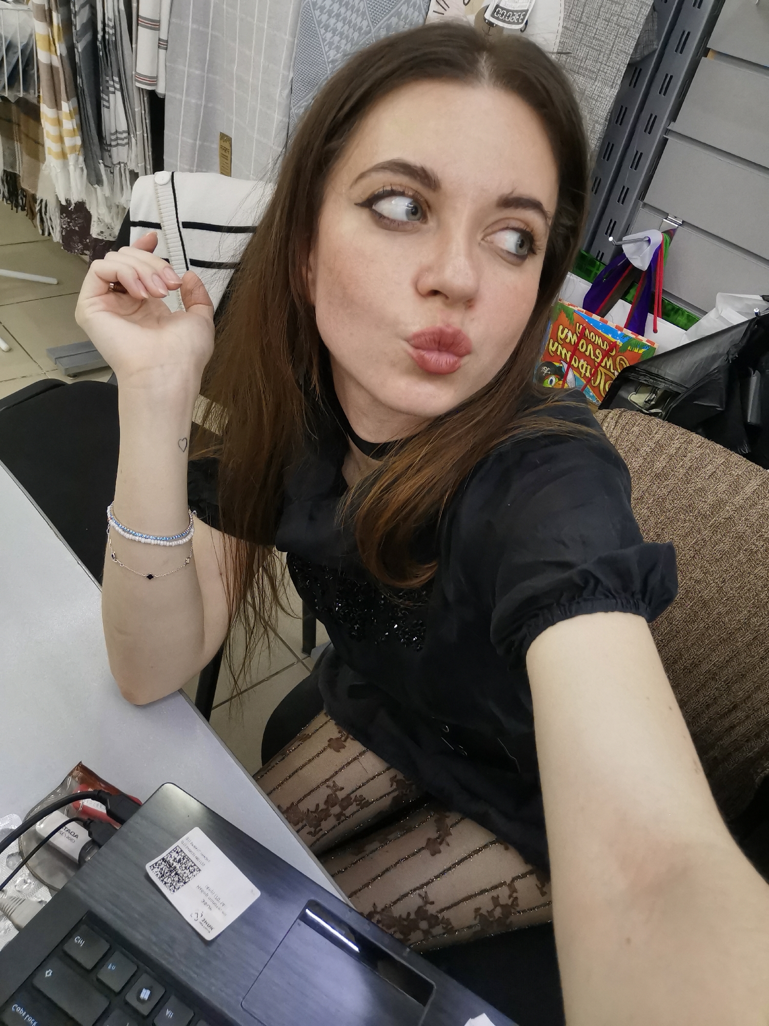 A medium shot depicts a fair-skinned woman with medium-brown hair, looking to her left with pursed lips. She is wearing a black choker, a black dress with short, puffed sleeves, and textured black tights. Her left arm rests on a white surface, adorned with two bracelets and a small heart tattoo on her wrist. She holds her right arm out to take the selfie.
The setting appears to be a store or business. To her left, merchandise hangs on racks, including scarves and clothing. Behind her, out of focus, are shelves with goods, and a shopping bag is visible. A laptop rests on the white surface in the foreground, obscuring details of the surrounding area. The location is unknown.