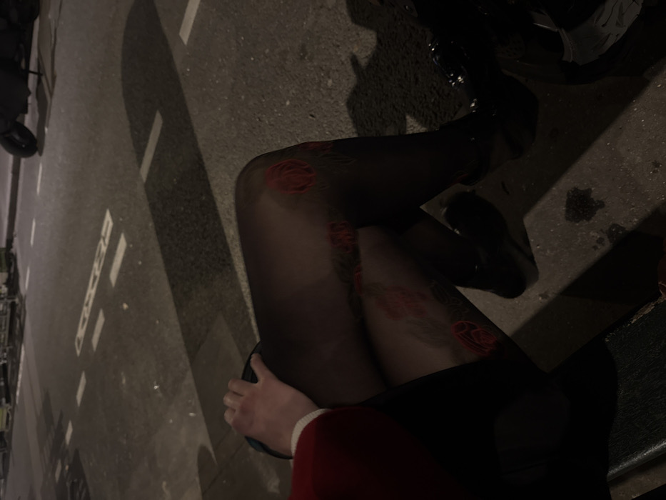 The image captures an outdoor nighttime scene, viewed from a low angle. The primary subject is the lower body of an individual, seated, with their legs extended. The person wears sheer black tights adorned with a repeating red floral, specifically rose, pattern. Dark shoes are visible on both feet. A hand with a light skin tone, covered by a red sleeve with a white cuff, rests on the individual's upper left thigh. The immediate foreground shows a dark, textured surface, likely a bench or seat.
The background consists of a dark asphalt street, illuminated by an unseen light source that casts long shadows. White dashed and solid road markings are present on the pavement. To the right side of the street, a dark scooter or motorcycle is partially visible, parked. The general environment is consistent with an urban street setting in Paris, France, characterized by paved roads, road markings, and parked urban vehicles like scooters. No specific landmarks are visible to pinpoint an exact location within the city.