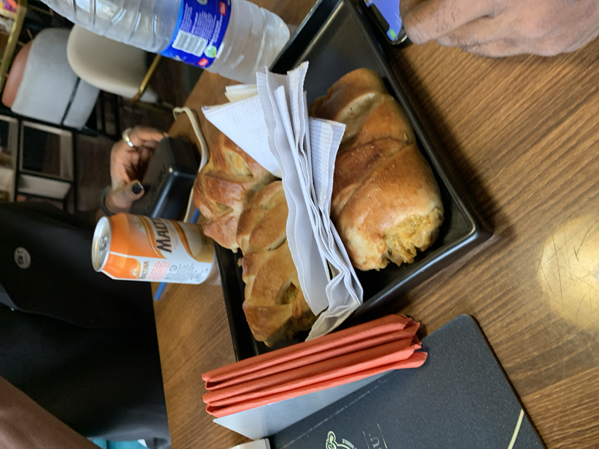 The scene depicts a table in Adankolo, Nigeria, with a partially eaten pastry on a black tray. The pastry is accompanied by white napkins and a stack of orange paper. A can of Malta drink and a water bottle are also present. Two individuals are partially visible; one person is seen reaching for something on the table, while the other is holding a phone. The table itself has a wooden finish, and a menu sits at the edge. The location is suggested by the presence of the Malta drink, which is commonly found in Nigeria.