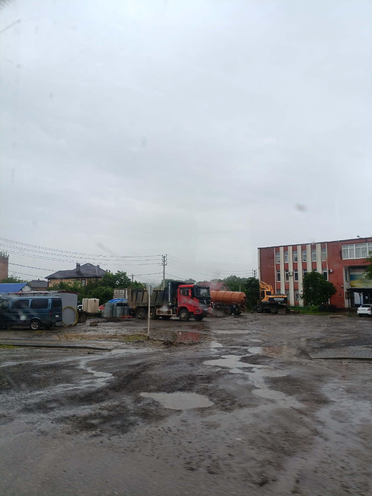 The image depicts a wet, possibly muddy, urban scene in Goryachiy Klyuch, Russia on an overcast day. The foreground is dominated by a large, damp, uneven area, likely a parking lot or open space. Visible in the middle ground are several work vehicles, including a dark-colored van, a dark gray dump truck, a red truck, an orange tank truck, and a yellow excavator. These suggest construction or maintenance activities are taking place. A two-story red brick building with white-framed windows is located on the right side of the image. Trees and greenery are scattered throughout the scene. In the background, the roofs of houses can be seen among the trees. Utility poles and wires are strung across the sky. A white car is parked in front of the red brick building.