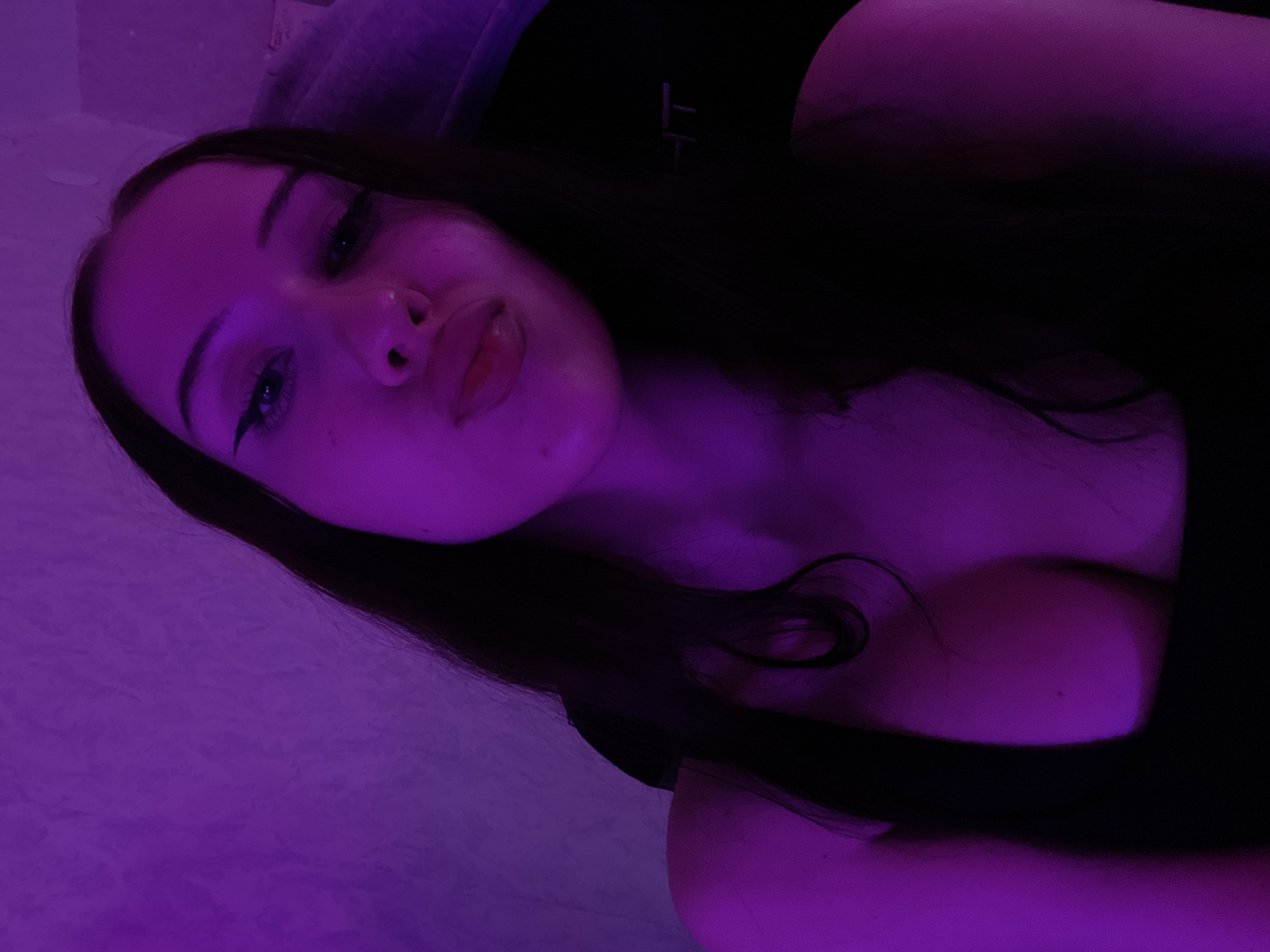 A young woman with long dark hair is captured in a selfie taken in what appears to be a dimly lit room, illuminated by purple lighting. Her facial features are prominent, with winged eyeliner accentuating her eyes and her lips pursed. She is wearing a dark-colored top that reveals a portion of her chest. The background is a textured wall, also bathed in the purple hue. A dark chair with a logo is visible behind her. There is no indication of the city of Votkinsk, Russia.