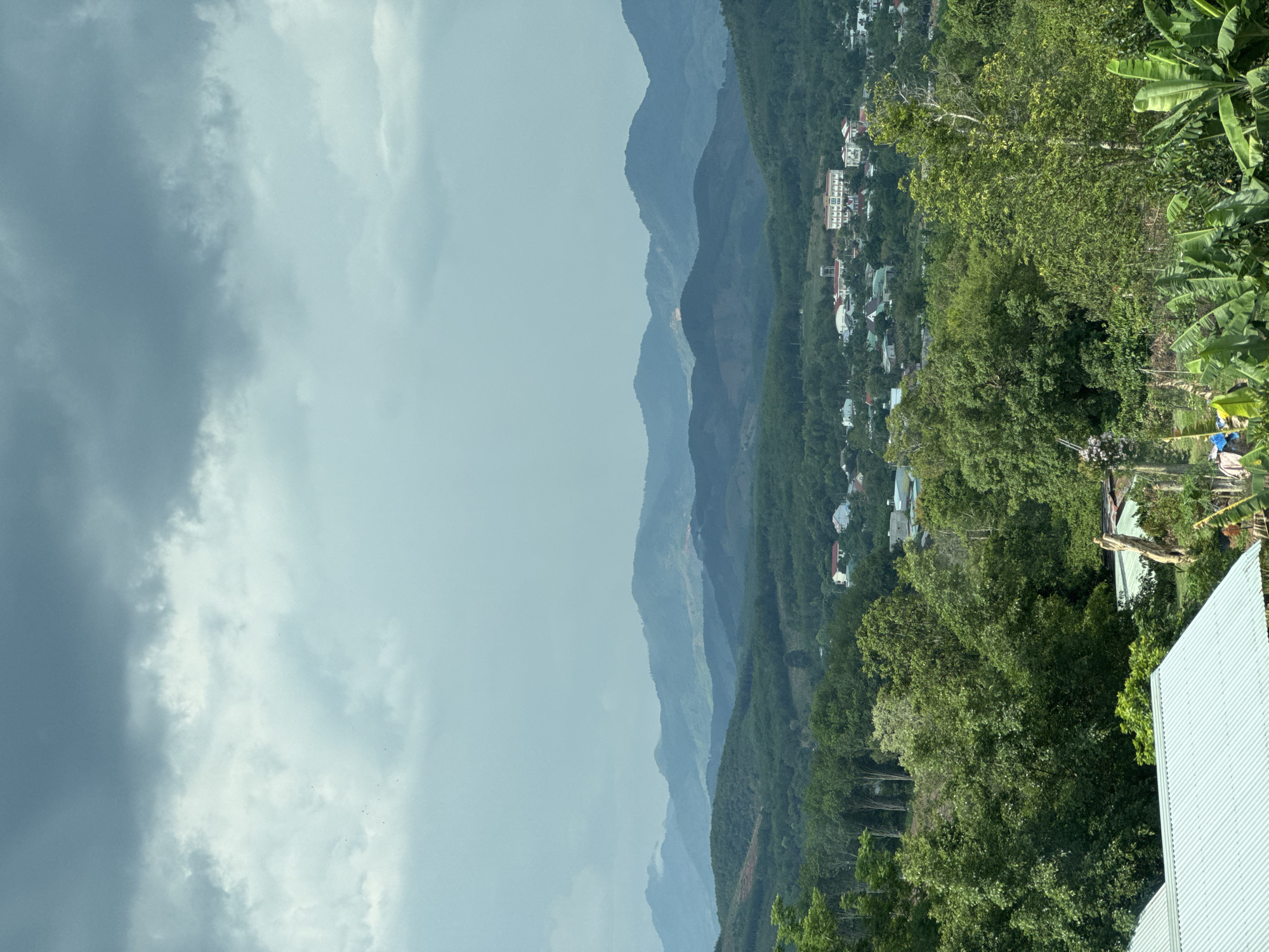 The image depicts a landscape in Tân Cảnh, Vietnam. The scene features a mountainous terrain veiled by a hazy atmosphere. A cluster of houses and buildings are interspersed within the dense vegetation on the mountainside. The sky is filled with an overcast layer of clouds. A rooftop is visible in the foreground. Banana trees are on the right side of the image.