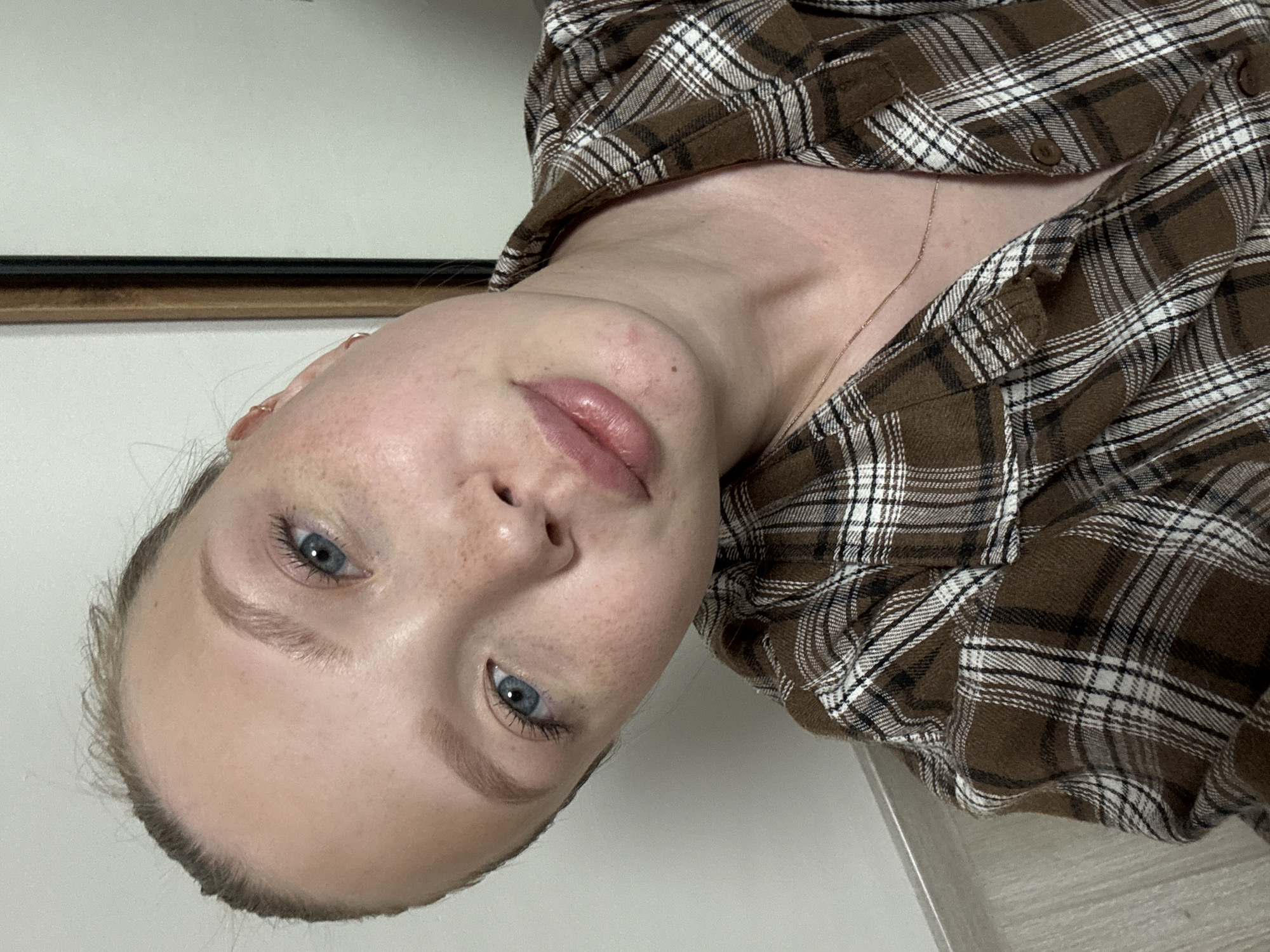 A young woman with blonde hair and blue eyes, wearing a brown and white plaid shirt, looks directly at the camera. She has freckles across her nose and cheeks. The background shows a white wall and a wooden floor, with a hint of a dark framed mirror on the right. The scene appears to be indoors, possibly in an apartment, in Chaikovsky, Russia.