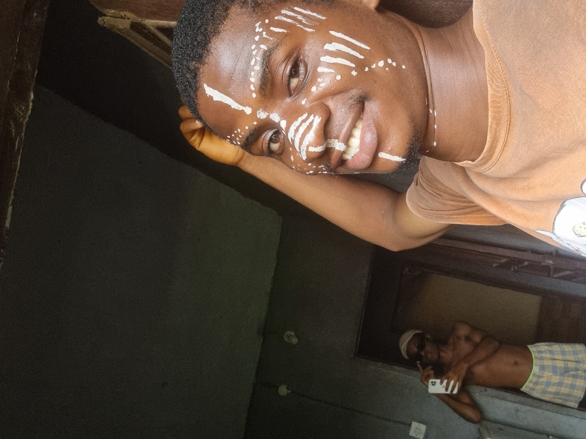This candid, rotated image, likely taken in Nkpor, Nigeria, features two young Black men in what appears to be an indoor or semi-enclosed setting.
The main subject, positioned prominently in the foreground, is a young man with white paint patterns adorning his face. These patterns consist of lines, dots, and stripes across his forehead, cheeks, nose, and chin. He is smiling broadly, looking directly at the camera with a joyful expression. He wears an orange or tan short-sleeved t-shirt and has short, dark curly hair. His head rests on his arm, suggesting a relaxed pose.
In the blurred background, another young Black man is visible, appearing to capture the moment on a white smartphone. He is shirtless, sporting a light-colored cap, dark sunglasses, and light-colored checkered shorts. He is leaning casually against a surface.
The setting appears to be a room or corridor with dark walls. A wooden beam or structure is partially visible in the upper left corner. The lighting suggests daytime, possibly from an unseen window or open doorway, illuminating the main subject brightly while the background remains somewhat dim. No visible text is present in the image. The overall mood is informal and cheerful.