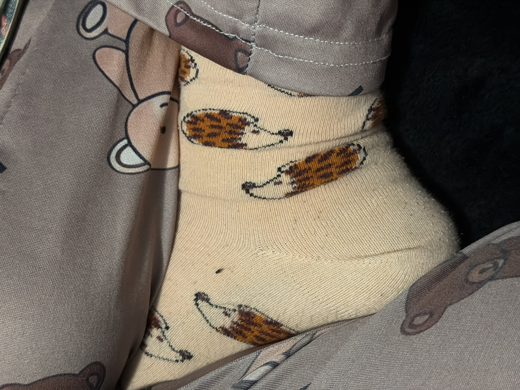 A foot wearing a beige sock with cartoon hedgehog patterns is visible between grey pajama pants with a cartoon bear print. The location is unknown and could not be deduced from the available information.