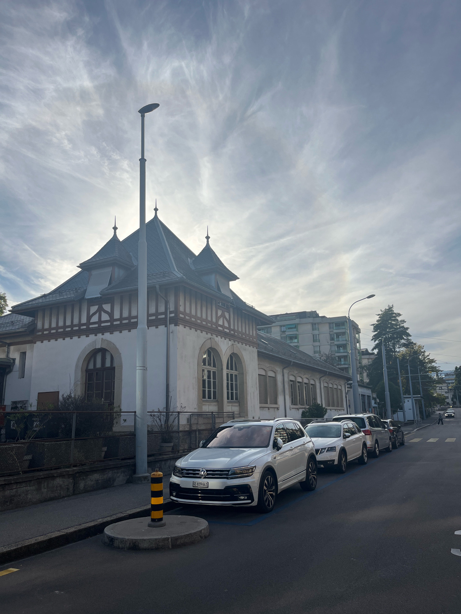 The image shows a street scene in Geneva, Switzerland, on a partially cloudy day. A white Volkswagen Tiguan is parked on the right side of the street, in front of a row of other parked cars. A yellow and black bollard is located on a circular concrete island in the foreground. Behind the bollard, a sidewalk runs alongside a building with a pitched roof and decorative wooden beams. Further down the street, there are other buildings, trees, and streetlights. A person is visible walking across the street in the distance, and car headlights are on. The sky has wispy clouds and a faint halo effect is visible.