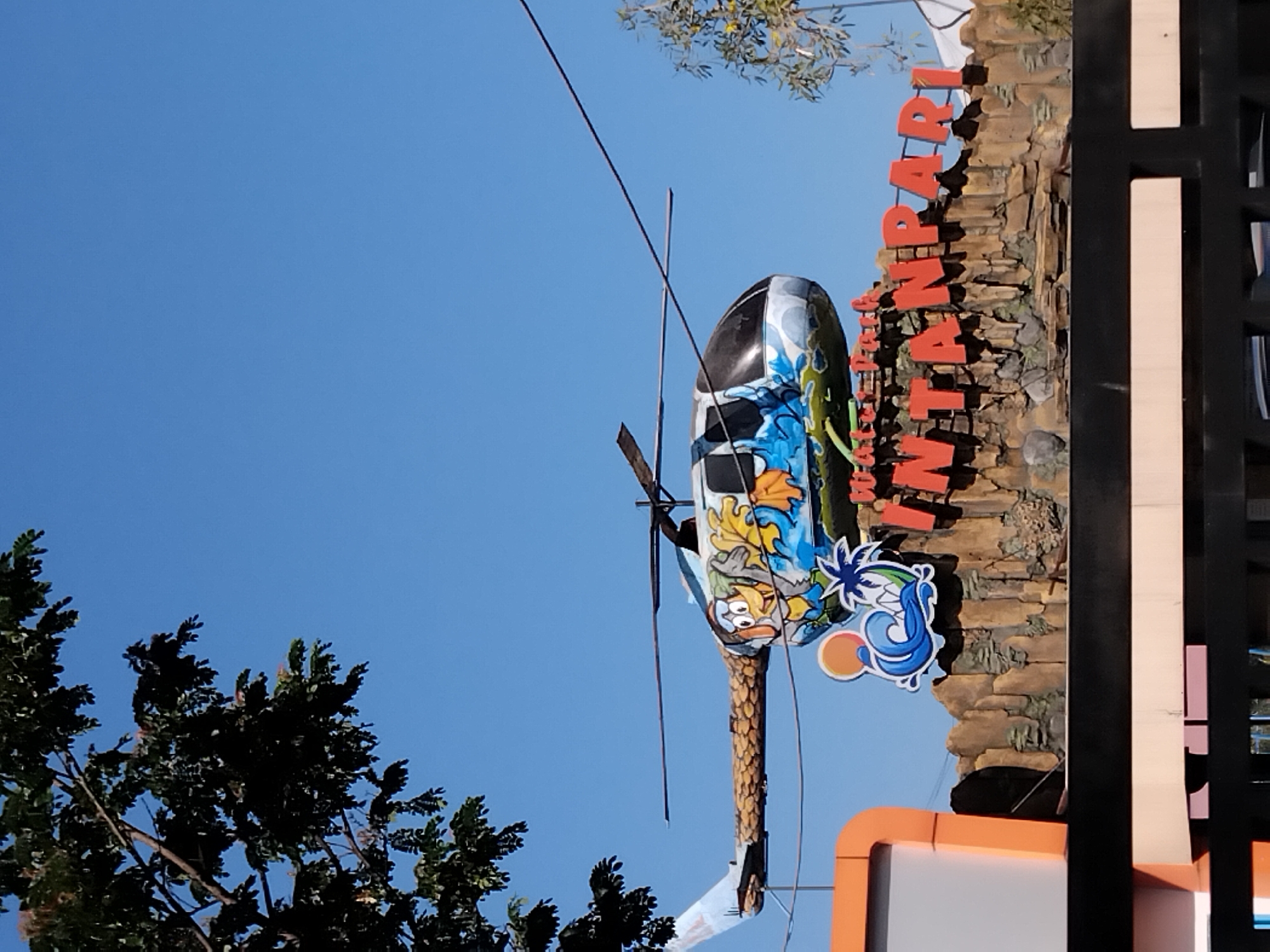 The image shows a vibrant outdoor scene under a clear blue sky, likely at a tourist attraction. Dominating the frame is a large helicopter prop, painted with cartoonish characters. This prop is mounted on a faux rock formation that serves as a sign. Large, red, three-dimensional letters spell out "INTANPARI," indicating the name of the place. Below the helicopter prop and to the left, there is a colorful graphic featuring a wave, a palm tree, and a sun, further suggesting a water park or similar recreational facility. To the far left of the image, an orange and white structure, possibly part of the entrance or a building, is visible. In the foreground, a black metal fence obstructs the view slightly. Trees with dark green foliage are seen in the upper left and upper right portions of the image, with some branches extending into the frame. The overall impression is that of a lively and family-friendly destination. This scene is located in Suruh, Indonesia.