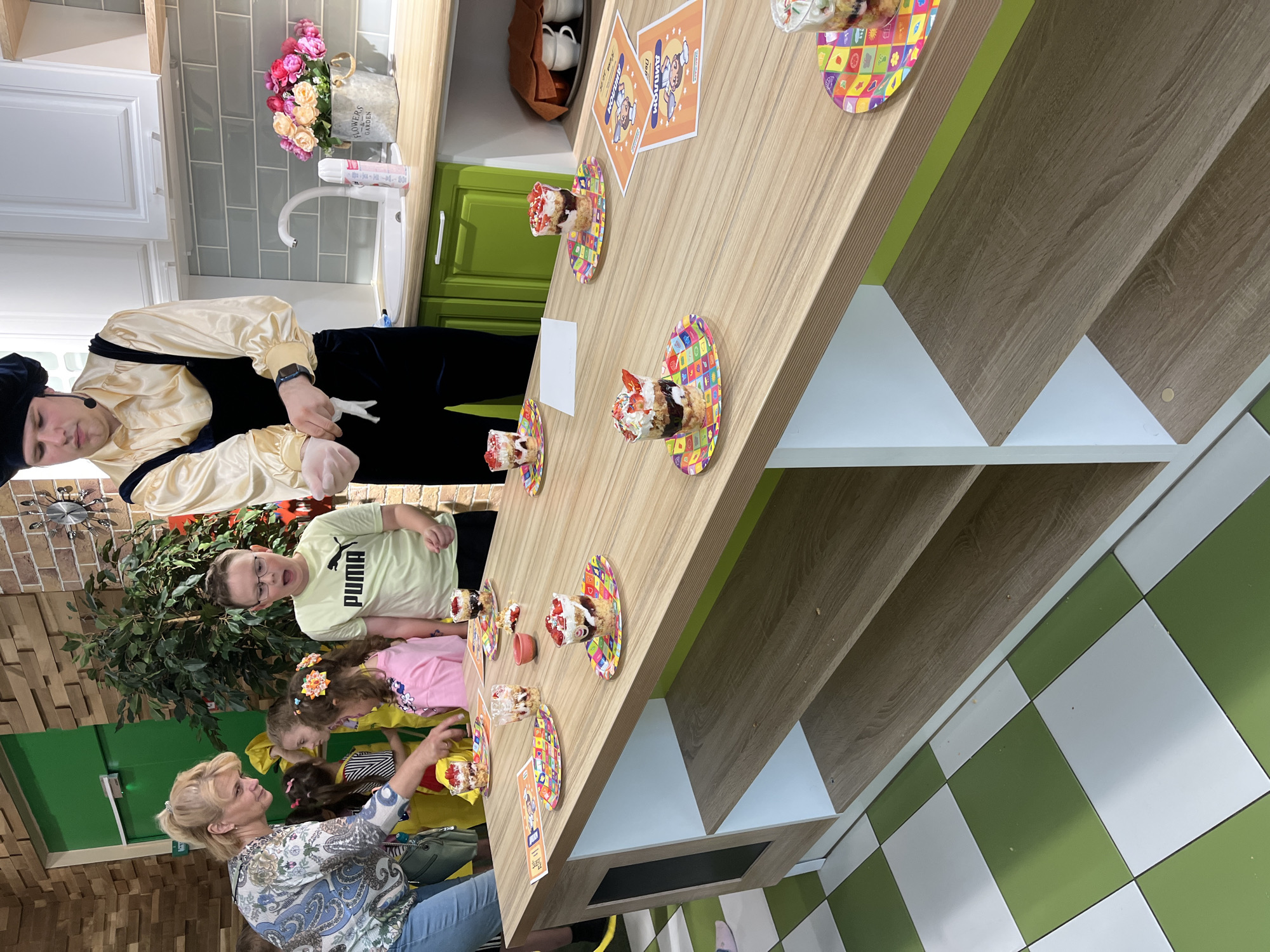 The image captures a children's party or cooking class scene. There are several children and two adults present, likely in a room with a kitchen setup. A man dressed in a period or themed costume stands at the head of a wooden table. The table is set with cupcakes on decorative plates, and promotional materials related to an event named "Am-nom" are on the table. The children, including one boy wearing a "PUMA" shirt, are standing around the table, appearing to be engaged in the activity. An adult woman, possibly an instructor, is helping them. The environment includes kitchen cabinets, a sink, and a floral arrangement. The floor has a checkered pattern of green and white tiles.