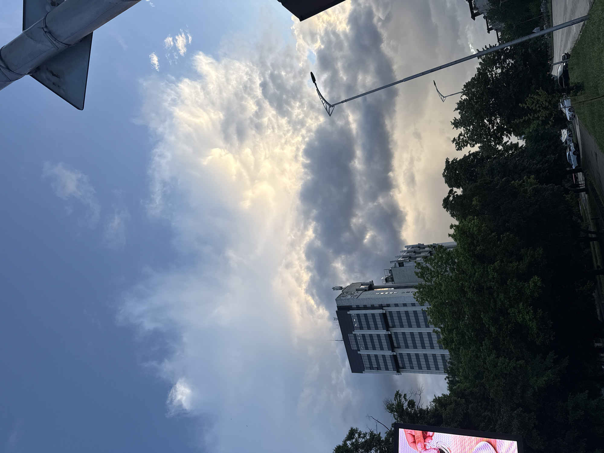 The image shows an upward angle view of a cityscape in Plovdiv, Bulgaria. The sky is filled with clouds, ranging from light blue to grey and dark shades, and suggests an overcast day. The sun is behind the clouds. A tall, modern building is centered on the right. There are large green trees on the right side of the building. On the extreme right of the image, there is a line of parked cars, behind which is a road. A streetlight appears near the top of the frame. On the left, there is a traffic sign pole. A portion of an electronic screen is visible.