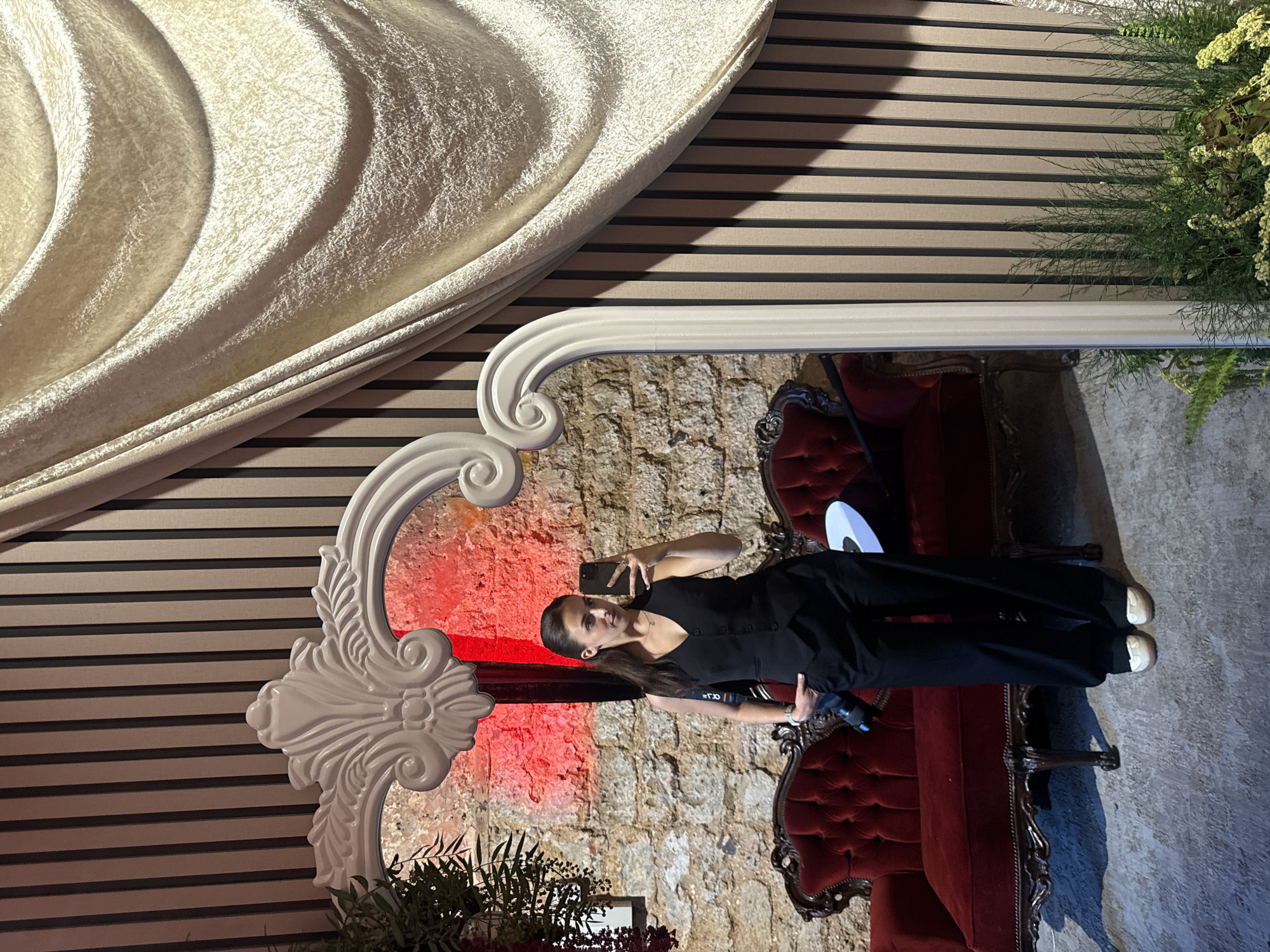 The image shows a person reflected in a mirror, seemingly taking a selfie. The person has dark hair and is wearing a black outfit. They are holding a phone in their hand.
The setting appears to be a decorated event space. The mirror has an ornate white frame. Behind the person in the mirror is a textured wall, possibly brick or stone, illuminated with red light. Two red antique armchairs are positioned on either side of the person. There are also floral arrangements visible, both at the bottom of the frame and to the right of the mirror.
The background behind the mirror frame consists of a beige and brown horizontally striped wall. A draped cream-colored fabric adorns the top-left corner of the scene, while additional greenery is present in the top-right corner.
It is not possible to confirm the location in Tel-Aviv, Israel, or any other city based on the visual information provided.