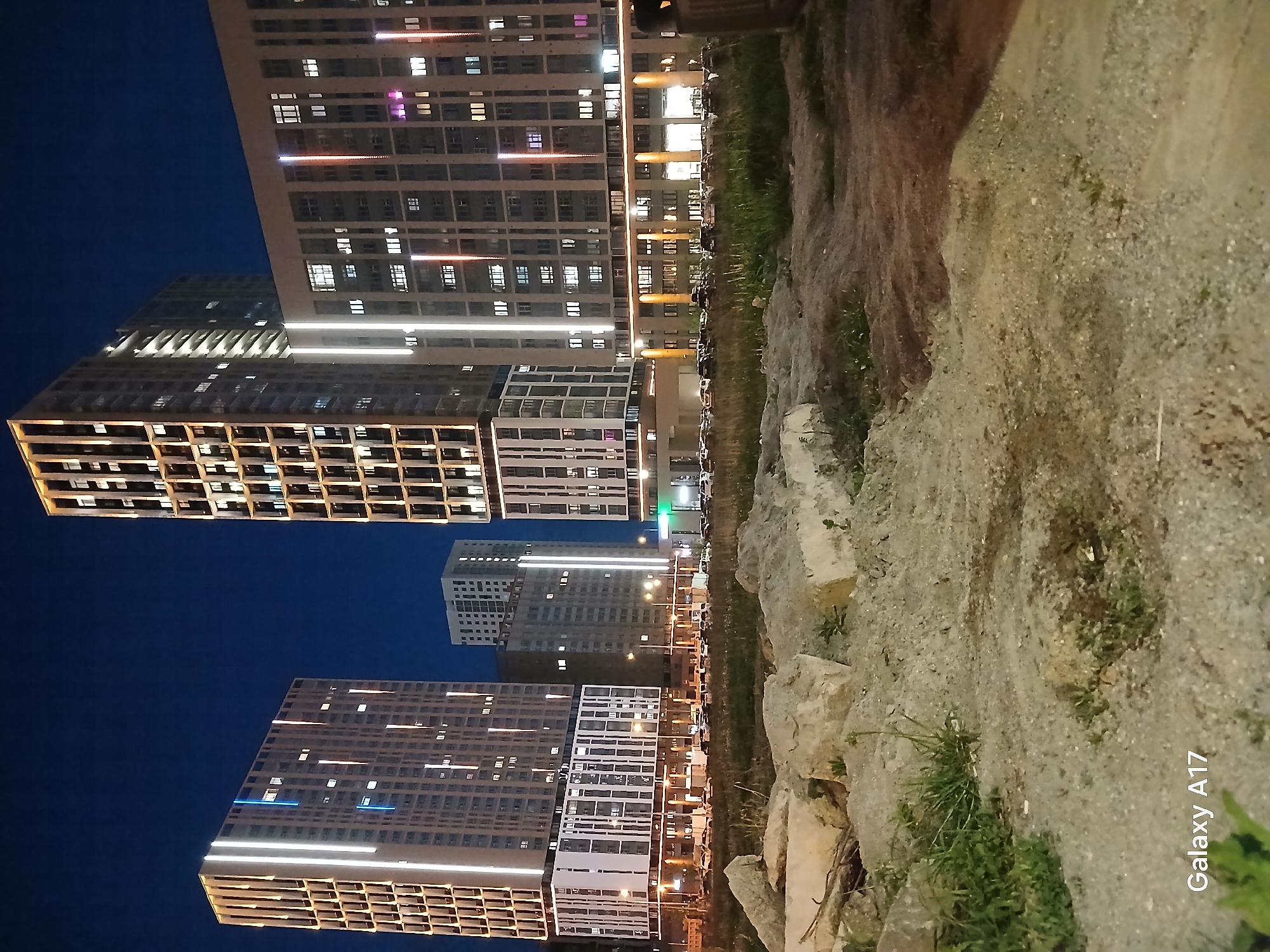 The image depicts several high-rise apartment buildings at night. It was taken in Yekaterinburg, Russia, as inferred from the Galaxy A17 watermark on the lower right of the image. The foreground shows a dirt path or construction area with rocks and grass. The apartment buildings are modern, with numerous lit windows arranged in a grid-like pattern. The night sky is a deep blue. The lighting suggests it is nighttime, with building lights illuminating the surrounding area. There is no presence of people in the image.