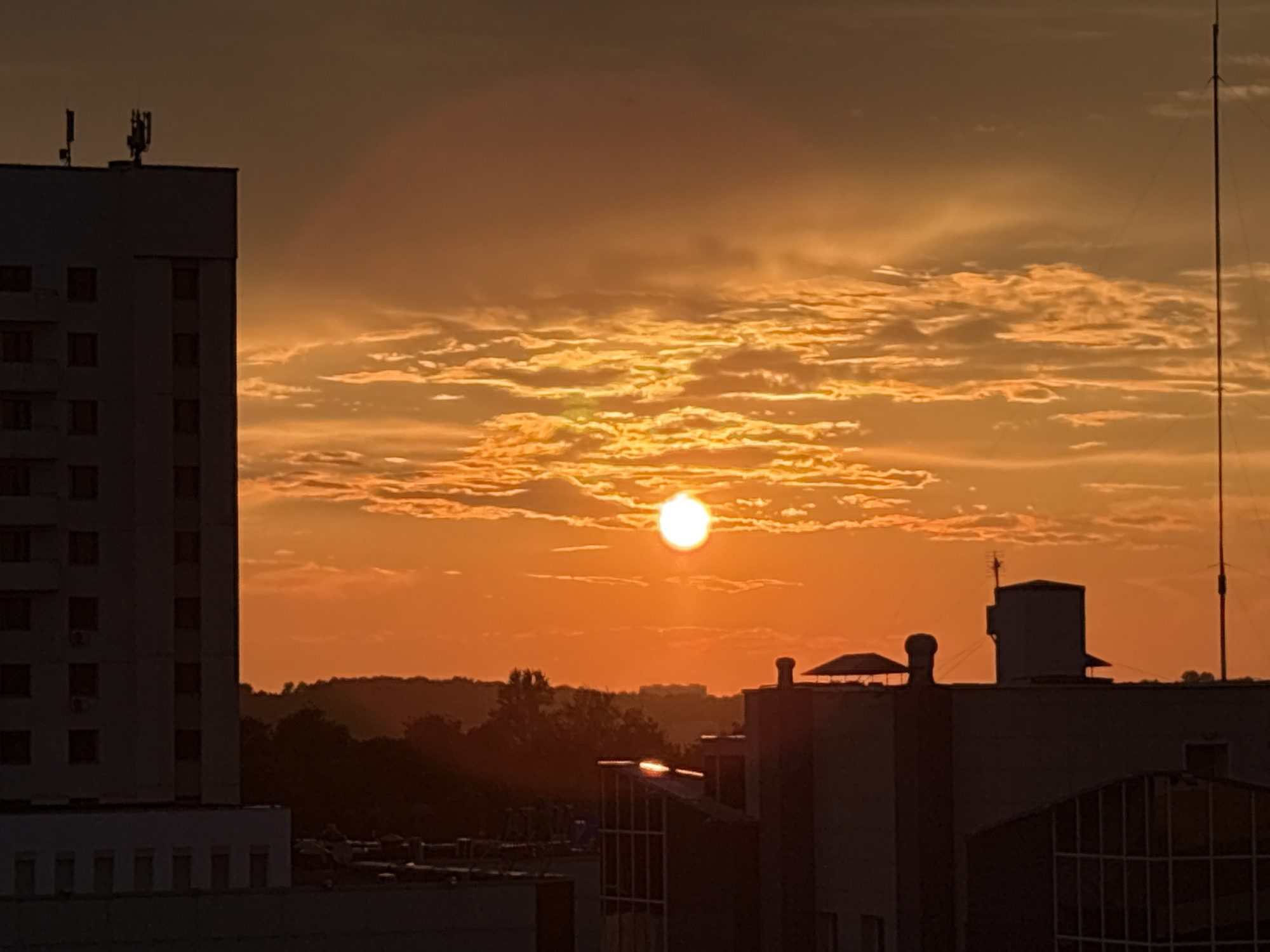 This image captures a sunset over an urban landscape, likely in Vitsebsk, Belarus. The sky is awash in vibrant shades of orange and yellow, with dramatic clouds catching the sun's rays. The sun itself is a bright, luminous orb, partially obscured by clouds, casting a warm glow over the scene.
Dominating the left side of the frame is a tall, multi-story building, silhouetted against the colorful sky. Its windows are dark rectangles, and the top of the building features telecommunications equipment, including antennae. In the foreground and mid-ground, other buildings are visible, also largely in silhouette, with some featuring rooftop structures like vents and what appear to be small shelters. A tall, thin radio tower stands on the right side of the image.
The setting appears to be late evening or early morning, given the intense colors of the sunset. The weather seems clear, allowing for a spectacular display of the sun. The presence of buildings and infrastructure suggests an urban or semi-urban environment. The overall mood evoked by the image is one of calm and beauty, with the natural spectacle of the sunset contrasting with the man-made structures. There is no visible text in the image.