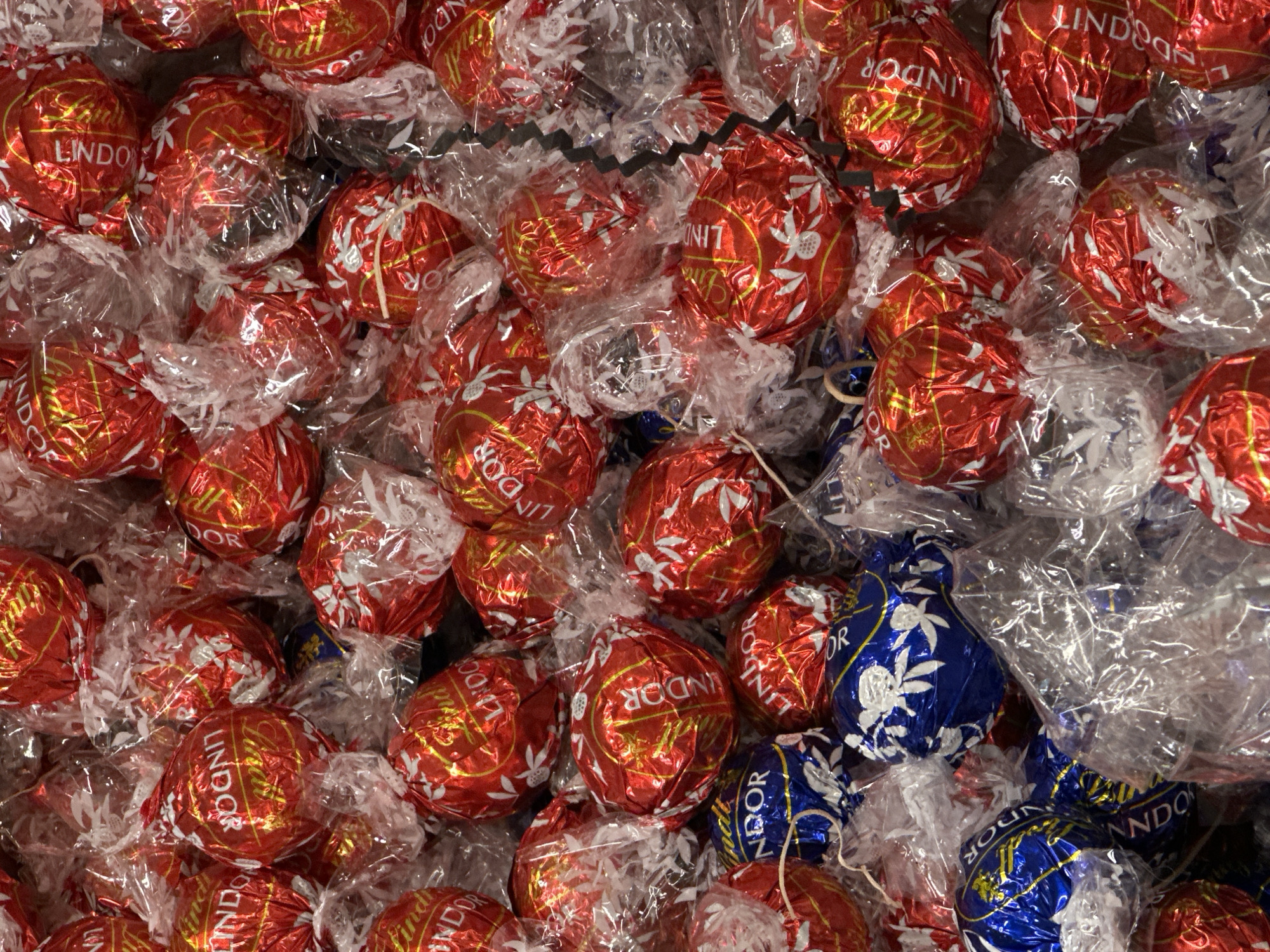The image shows a close-up, high-angle view of a collection of Lindor chocolate truffles. The majority of the truffles are wrapped in red foil with white floral designs and the word "Lindor" printed on them. A smaller number of truffles are wrapped in blue foil, also with white floral designs and "Lindor" branding. The truffles are individually wrapped in clear cellophane, which is crumpled and adds texture to the scene. The truffles are densely packed together, obscuring any background details. The image does not provide any information about the location in Vancouver, Canada or any additional context.