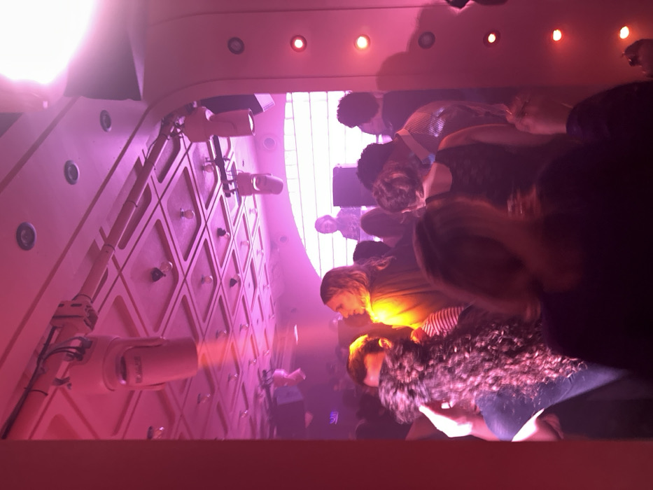 This image captures an indoor, crowded venue, likely a club or bar, located in Paris, France. The scene is dominated by strong pink and magenta artificial lighting, creating a vibrant, atmospheric ambiance enhanced by a visible haze. The ornate, coffered ceiling features numerous recessed light bulbs and track lighting fixtures, from which a pale, cylindrical speaker labeled "MONCEAU" is suspended. On the right, a curved architectural element is lined with a vertical series of small, circular red accent lights. In the background, an elevated platform holds a DJ, positioned behind mixing equipment and illuminated by bright white light emanating from a large, arched, grid-patterned window or light panel. A dense crowd of individuals, predominantly adults, fills the space. Notable figures include a person with long, light-colored hair wearing a yellow top in the mid-ground, and a person with dark, curly hair in the left foreground. The collective presence of people suggests social interaction, dancing, or live music engagement.