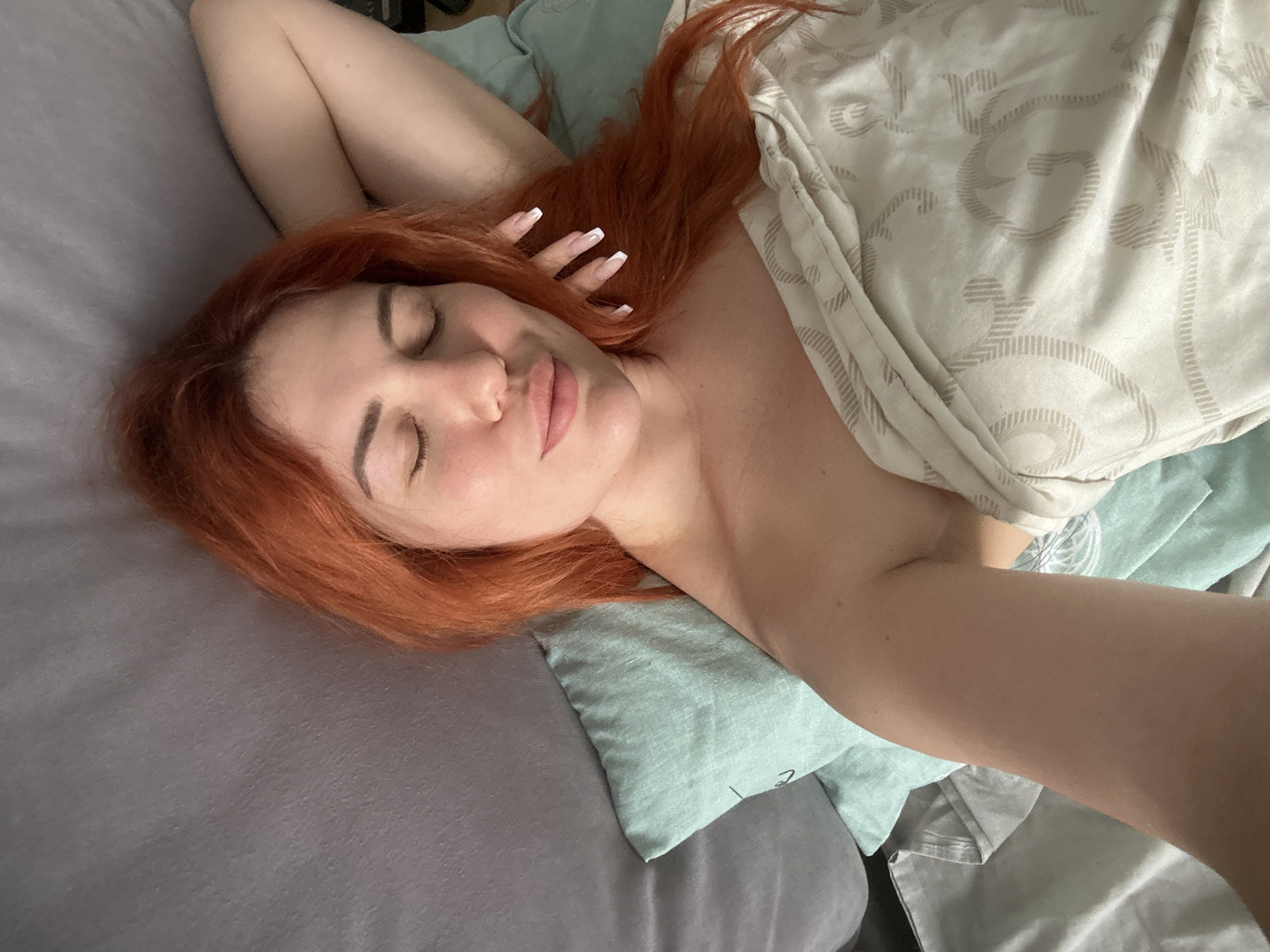 In a close-up shot taken from above, a person with vibrant red hair rests in bed, eyes closed, suggesting they are asleep or deeply relaxed. Their head is propped on a light blue pillow, with a gray surface, possibly a headboard or another pillow, behind them. The person is partially covered by a beige blanket adorned with a swirling pattern. One arm is raised and bent behind their head, with manicured fingernails visible. The lighting is soft, casting a gentle glow on their face and arm, highlighting their features. The scene appears to be in a domestic setting, likely a bedroom in Odesa, Ukraine.