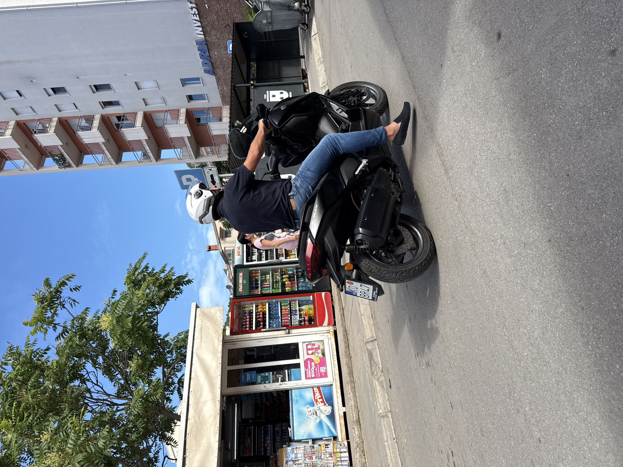 A man wearing a white helmet, dark polo shirt, blue jeans, and black loafers is riding a black scooter. He is on a paved street with a sidewalk to his left. To the left of the scooter is a storefront with a white awning, displaying various products. Beyond the storefront, a tall, modern apartment building with multiple balconies is visible. The building has "ADRIA INVEST" signage. The sky is blue with some clouds. The scooter has a license plate that reads "KO BR47". The scene appears to be in the city of Budva, Montenegro.