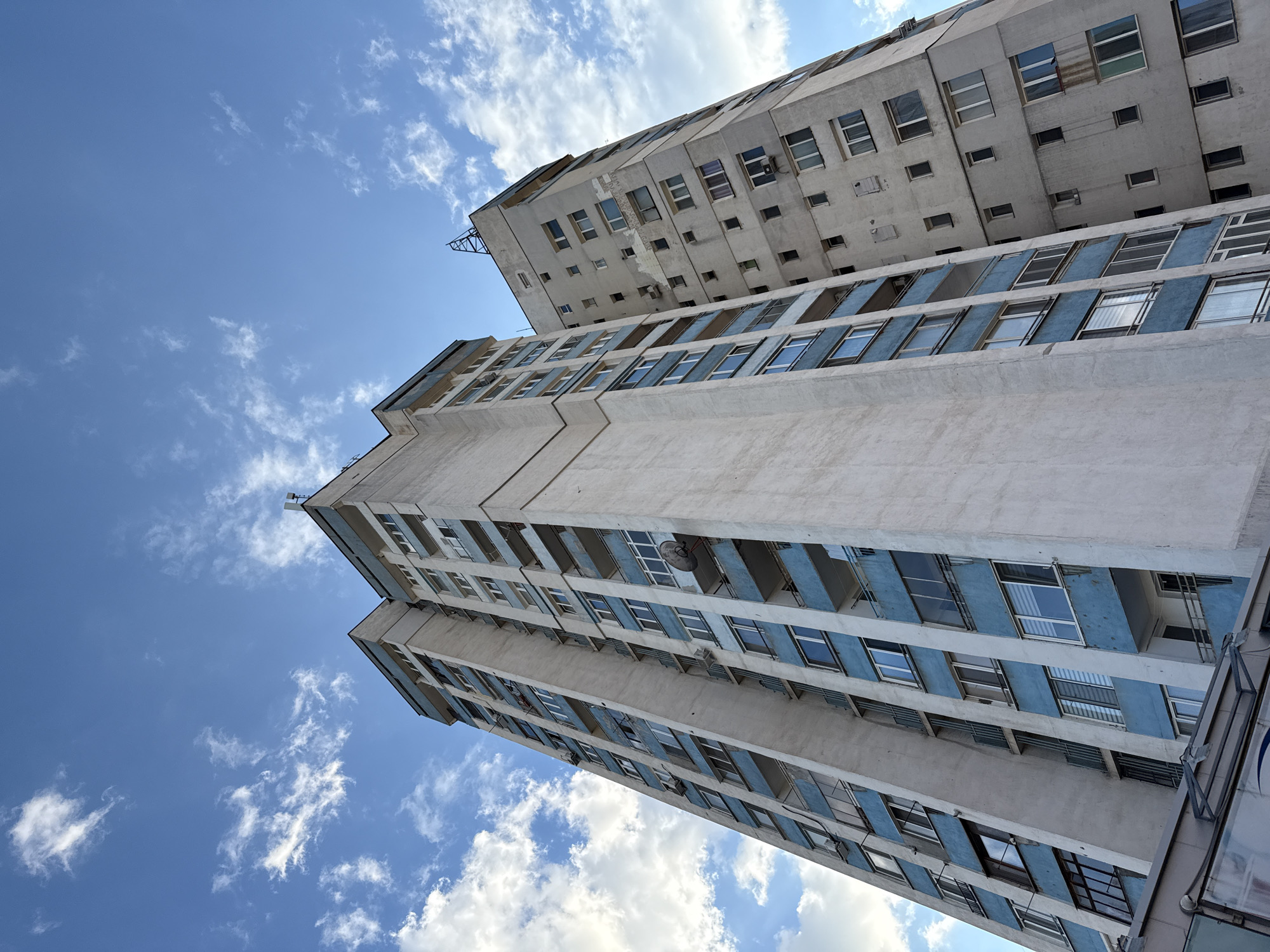 The image shows a low-angle view of two tall apartment buildings in Sofia, Bulgaria, against a backdrop of a blue sky with scattered white clouds. The buildings appear to be of Soviet-era construction, characterized by their utilitarian design and concrete exterior. The lower building features rows of windows with blue accents, while the upper building has a more varied arrangement of windows. Both buildings show signs of age and wear. The perspective emphasizes the height and verticality of the structures. No people are visible in the image.