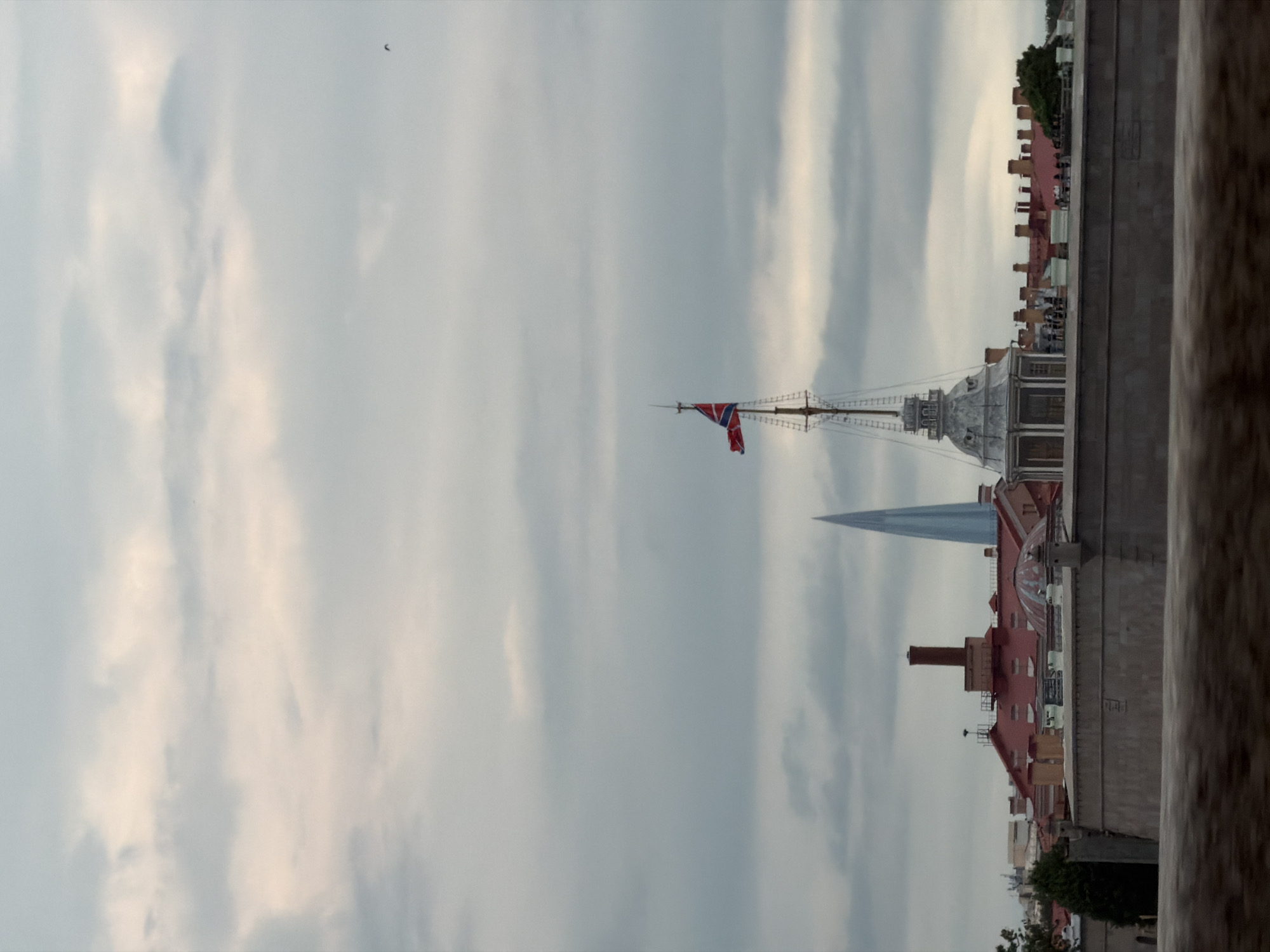 The image captures a cloudy sky over a cityscape in Russia. In the foreground, a stone wall or embankment runs horizontally across the lower portion of the frame. Behind it, a collection of buildings with red roofs and chimneys are visible. A prominent structure with a dome and a tall, spire-like tower stands out, with a flag flying from its mast. To the left of this structure, a tall, slender, pointed building, possibly a skyscraper, pierces the sky. The overall impression is of an urban landscape under an overcast sky.