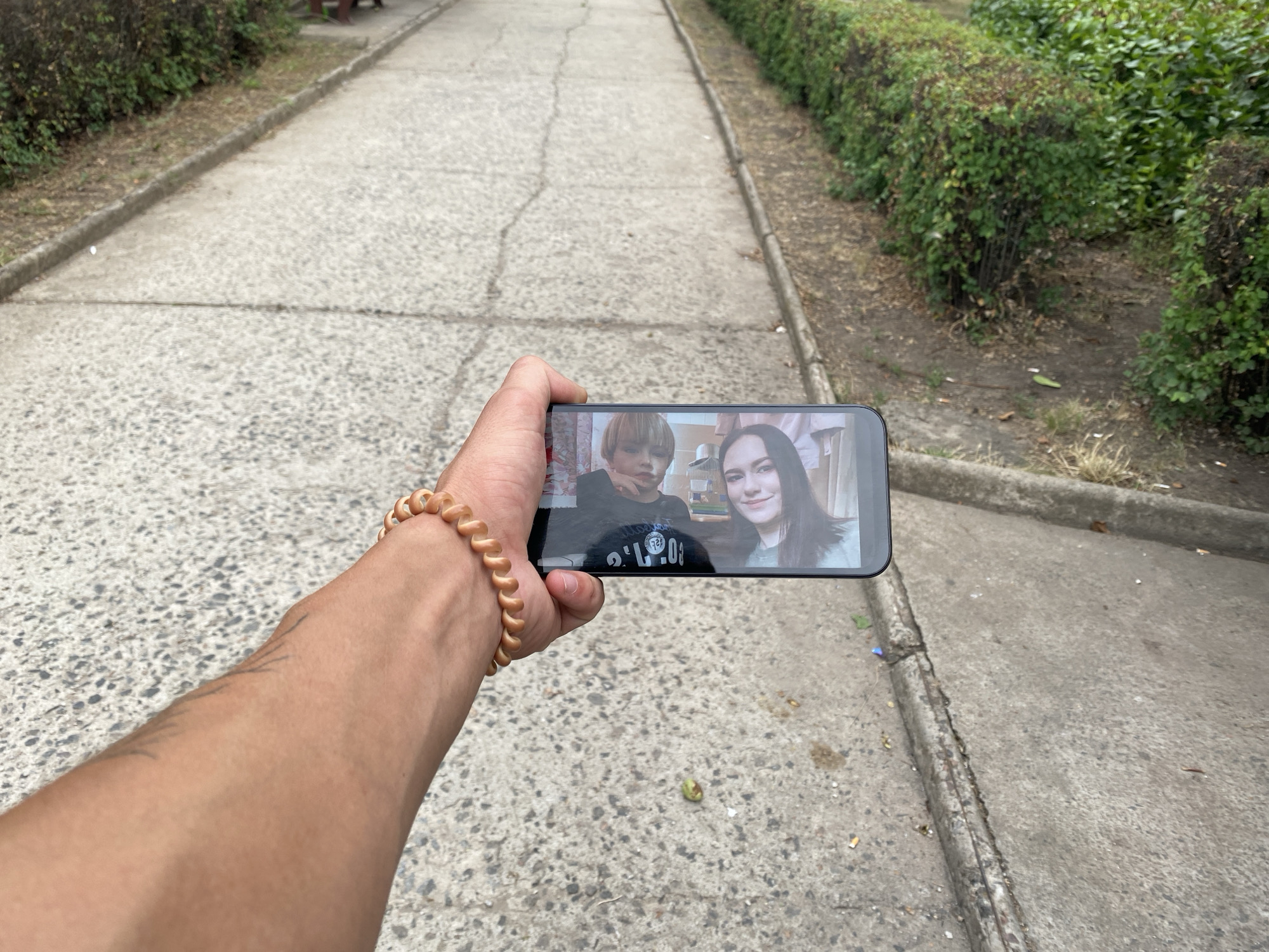 A first-person perspective shows a person's arm and hand holding a smartphone, displaying a video call. The smartphone screen shows two people: a young boy with blonde hair on the left and a young woman with long dark hair on the right. The arm has a spiral-shaped bracelet made of light brown beads. The background is a paved path with a concrete curb and manicured green bushes lining one side. The path appears to be in a park or along a street in Pivdennoukrainsk, Ukraine, indicated by the presence of numerous cigarette butts scattered on the ground near the curb. The overall impression is of an outdoor, urban environment.