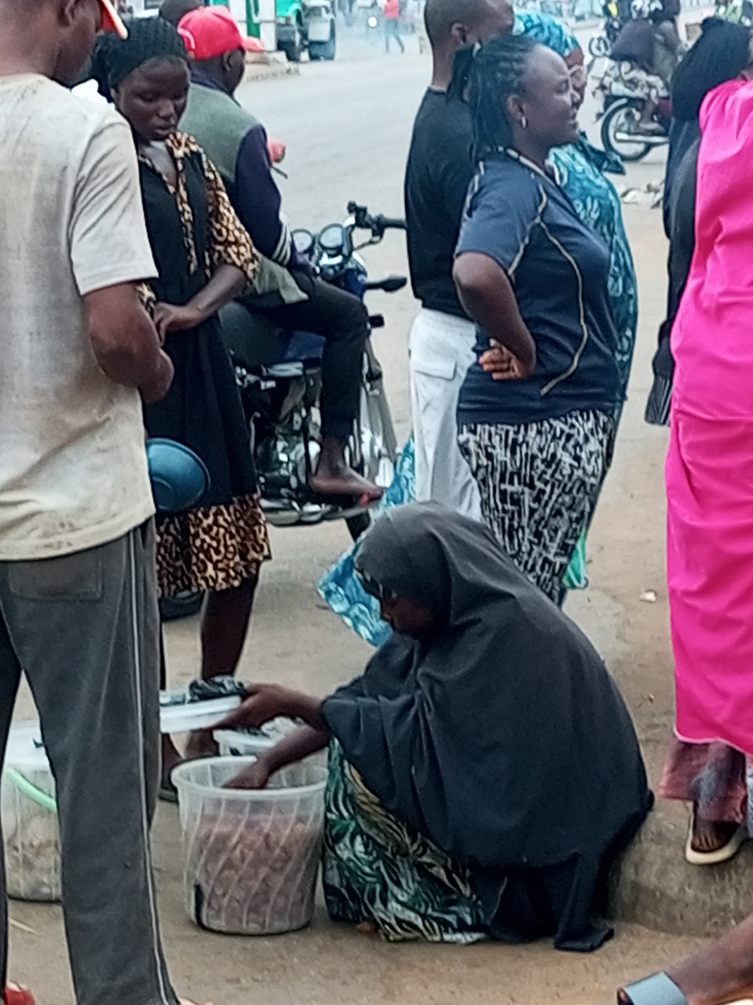 A group of people are gathered on a dusty street, likely in Gombe, Nigeria. In the foreground, a person wearing a black hijab and a patterned skirt kneels beside a large clear plastic bucket filled with a reddish-brown material. Their right hand is extended towards the bucket. To their left, another person in grey trousers and a white t-shirt stands. Behind the kneeling figure, a woman in a dark blue shirt and patterned pants stands with her hands on her hips. To her left, a woman in a black headscarf and a black and leopard print dress is seated on what appears to be a motorcycle. Further back, several other individuals are visible, some standing, others on motorcycles. A bright pink garment is visible on the far right side of the image. In the background, there are buildings and a glimpse of what might be a market or busy street. The overall scene suggests a street-side transaction or gathering in a bustling urban environment.