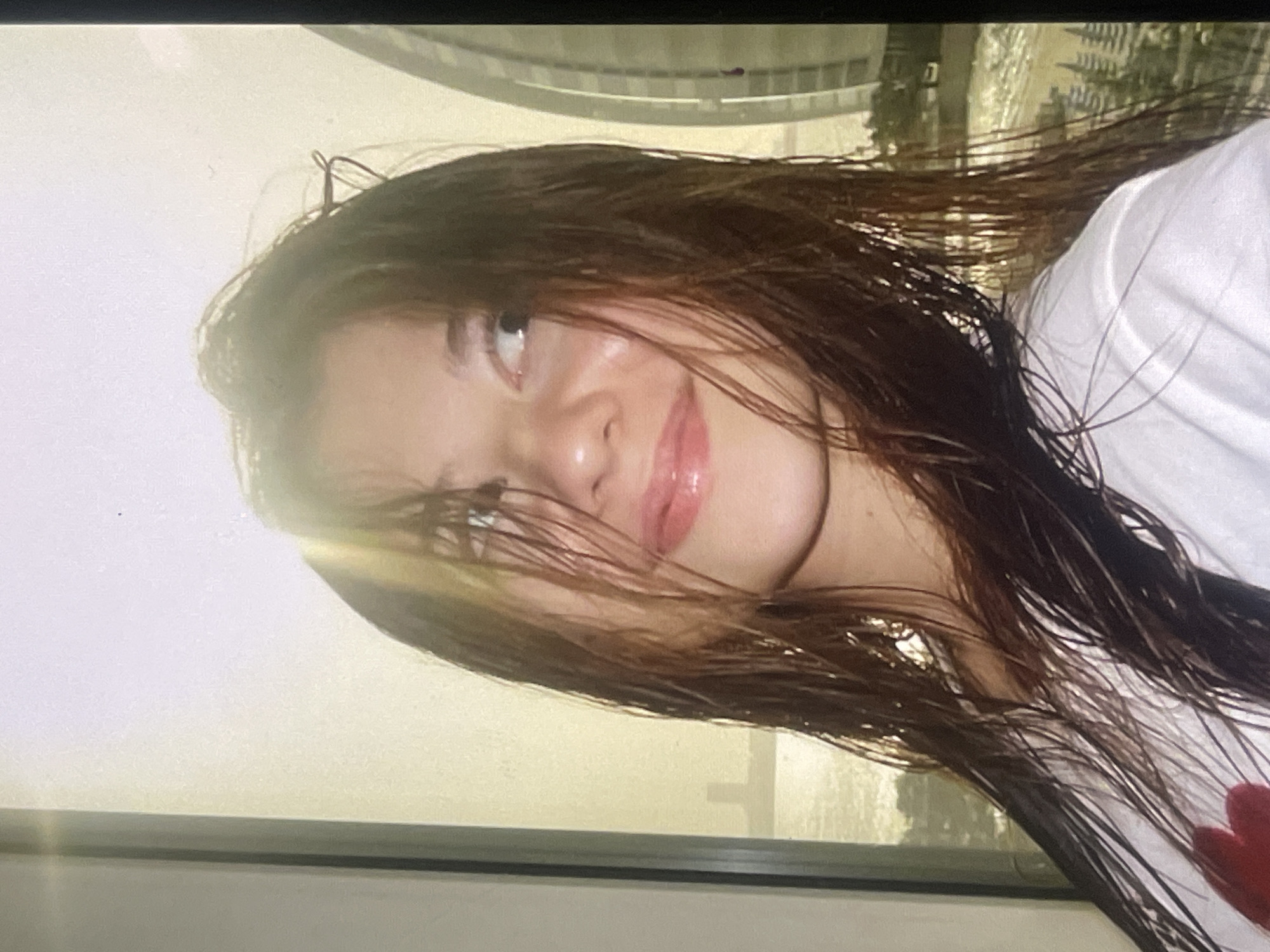 A young woman with long, dark, wet-looking hair smiles softly while looking upwards and to the left. She has glossy lips and appears to be in her late teens or early twenties. She is wearing a white t-shirt, with a glimpse of a red heart-shaped detail visible on the lower right side of her garment. The image is captured indoors, looking out through a window, and is slightly tilted.
Through the window, a bright, urban landscape is visible. On the upper left, a prominent, curved, modern architectural structure, possibly metallic or glass, dominates part of the background. Further to the right and in the mid-ground, other buildings and structures can be vaguely discerned, suggesting a cityscape. The lighting is bright, indicating daytime, with a strong light source creating a flare near her forehead and illuminating her face. A window frame or sill is visible along the bottom edge of the image. The overall mood is casual and pleasant. No text is visible.