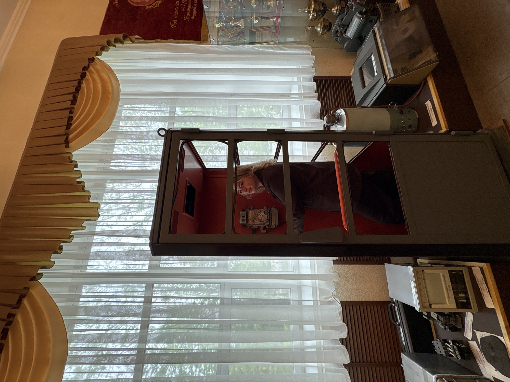 A person is inside a dark, box-like structure with small windows, possibly an old-fashioned photo booth or scientific instrument. The interior of the box is red. The person is looking towards the viewer. Outside the box is a window with white curtains. Behind the box is a display of items, including what appears to be awards and trophies in a glass case. In the background, there are reddish drapes. The scene appears to be indoors, in a museum. This image may be associated with the city of Birobidzhan, Russia.