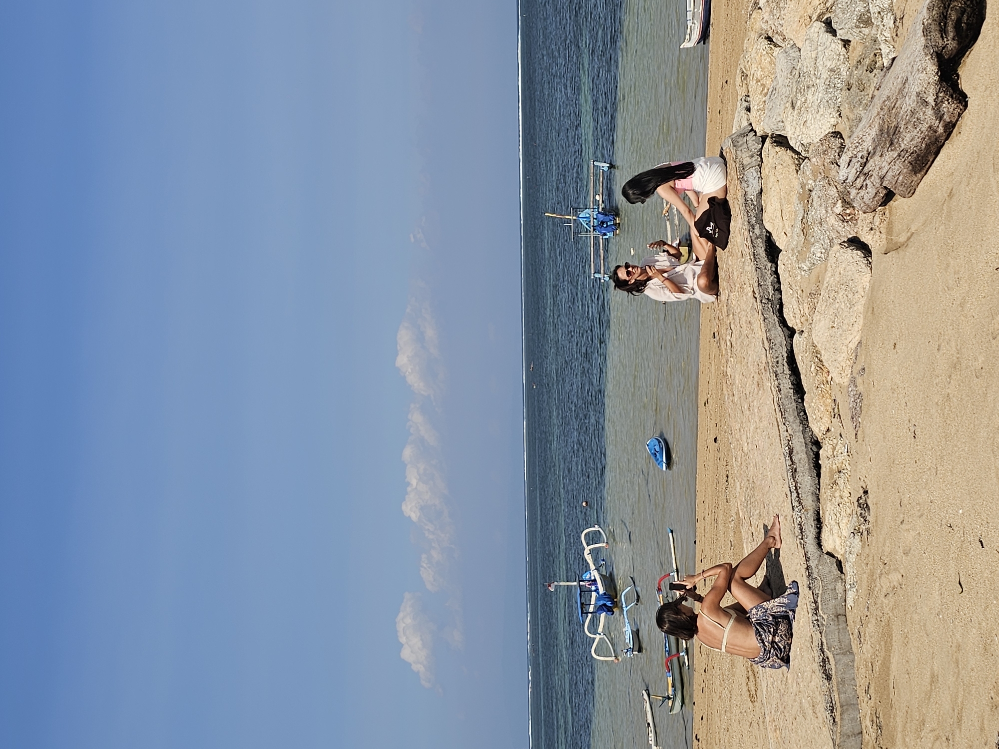 The image shows a tropical beach scene in Denpasar, Indonesia, with a bright blue sky and scattered white clouds. In the foreground, three individuals are present. On the left, a woman wearing a bikini top and a patterned sarong is sitting on the sand, facing away from the camera, and appears to be taking a photo with her phone. In the middle ground, two women are sitting on the sand near some rocks, facing each other. One is wearing a white shirt and sunglasses, and the other is wearing a pink tank top and white shorts. Both appear to be holding drinks and engaging in conversation. Several traditional Balinese fishing boats, known as jukung, are anchored in the shallow, clear water. One larger jukung with a blue canopy is positioned centrally, and other smaller boats are visible scattered across the water and shoreline. The beach itself is sandy, with some rocky outcrops and a large piece of driftwood in the lower right corner of the image. The ocean water is calm with a visible horizon line and distant landmass.