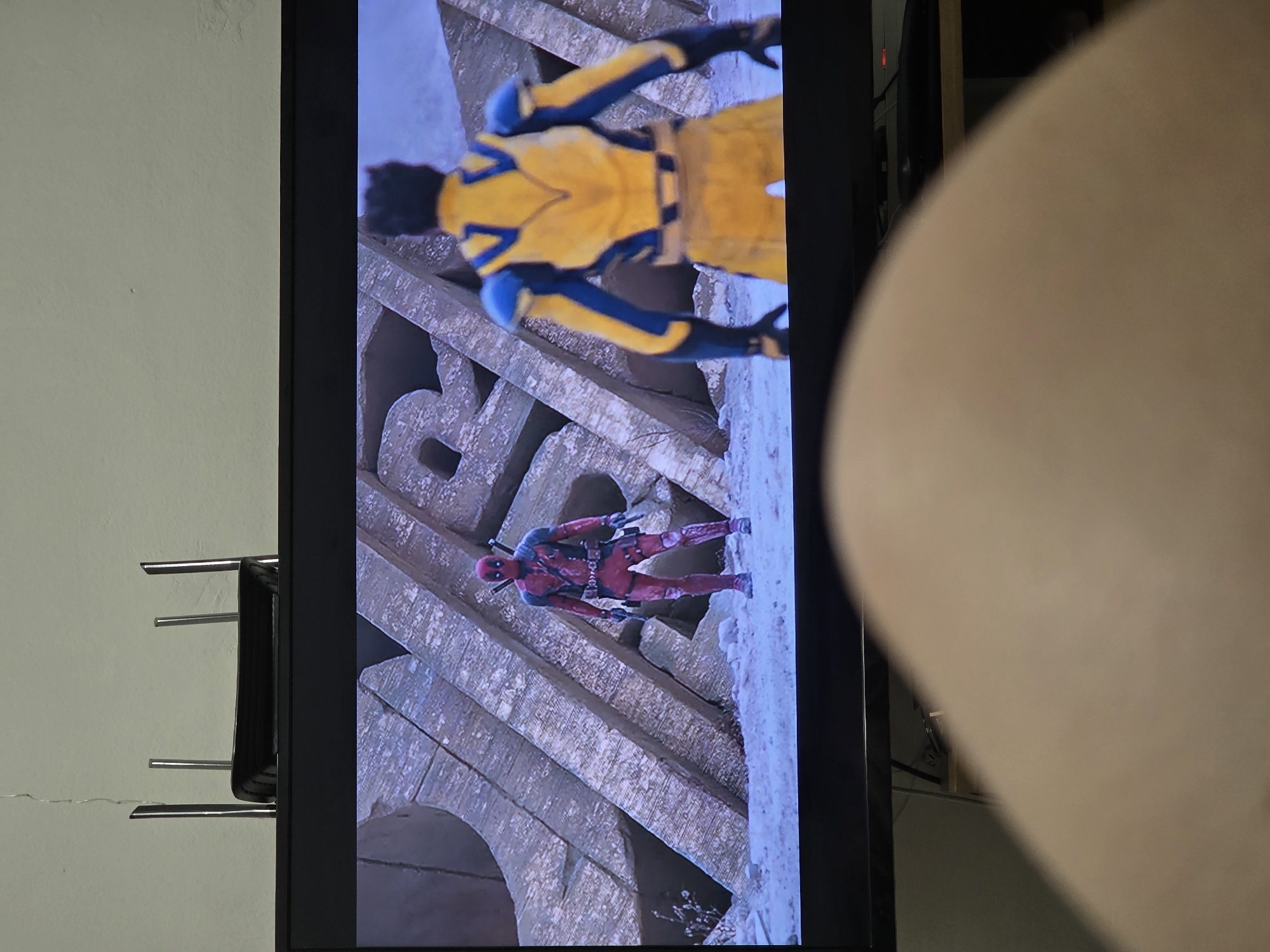 In the foreground, a person's thigh is blurred due to the close proximity to the camera, obscuring the lower portion of the image. Dominating the frame is a television screen displaying a scene from a movie. On the screen, two figures are visible. To the left, a character in a red and black suit, resembling Deadpool, stands with two swords sheathed on his back. To the right, another character, seen from behind, wears a yellow and blue costume, likely Wolverine. They are positioned in an outdoor environment with large, weathered stone structures and a snowy ground. Behind the television, a black router with multiple antennas is placed on top of the television. The background wall is a light color with visible cracks.