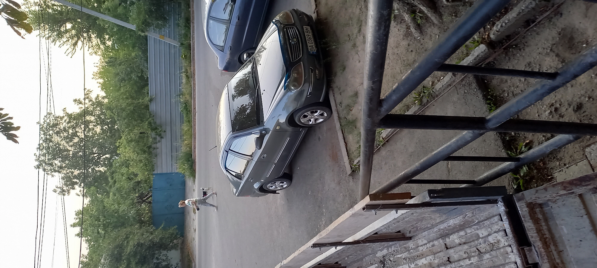 The image captures a street scene in Kropyvnytskyi, Ukraine. In the foreground, a rustic wooden structure with metal railings is visible, suggesting a construction site or a loading area. Beyond this, a grey sedan is parked on the asphalt, its license plate reading "BA 0369 BM". A second, darker sedan is partially visible behind it. To the left, a woman is walking a small dog on a leash on the sidewalk. The background features a line of trees and a corrugated metal fence. The sky is overcast, and the lighting suggests it is either early morning or late afternoon. The overall impression is that of an ordinary urban environment.