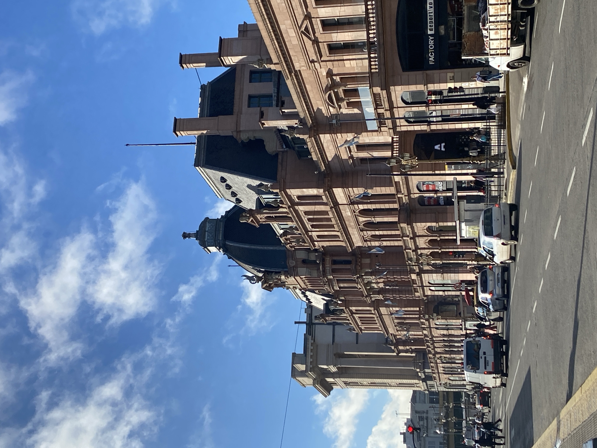 This is a daylight photograph of a bustling urban street scene, likely in Buenos Aires, Argentina, given the location context and architectural style. The image is oriented vertically, with a bright blue sky and scattered white clouds at the top.
The left side of the frame is dominated by a grand, light-colored building featuring ornate classical architecture, multiple stories, numerous windows, and balconies. A prominent dark-domed roof, topped with decorative elements and statuary, adds to its distinctive appearance. Gargoyle-like figures are visible beneath the roofline in places, and several chimneys extend upwards.
The wide street occupies the right and lower portions of the image, marked by white dashed lines indicating lanes and a yellow curb. Various vehicles, including cars, several white vans, and a white pickup truck with an open bed, are present on the road, suggesting active traffic or parked vehicles. A red traffic light can be seen in the distance on the lower left.
A few pedestrians are visible on the sidewalks next to the building. The strong sunlight casts distinct shadows, indicating a clear, bright day, likely late morning or early afternoon.
Visible text on storefront signs on the building includes "FACTORY deportivo" on a black awning, a stylized "A" logo on another sign, and "HEAD" on a sign closer to the street level. The scene depicts a vibrant city environment, blending historic architecture with contemporary street activity.