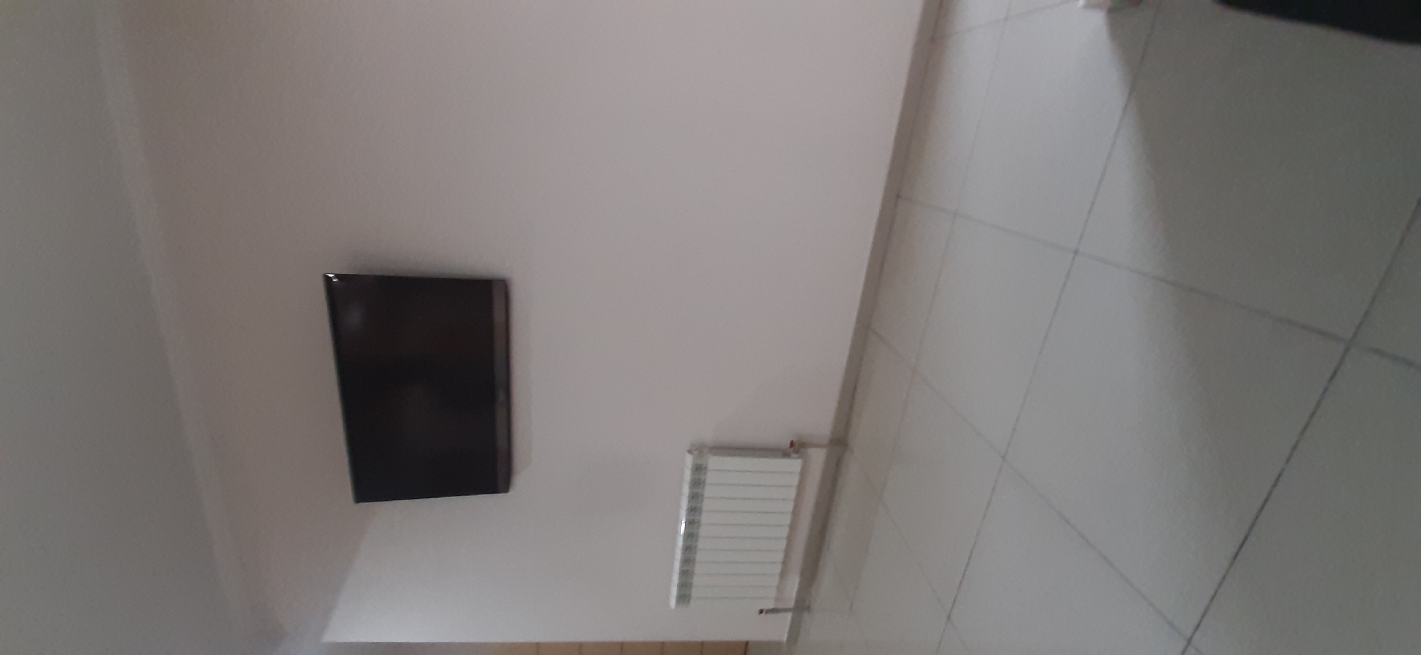 This image shows an interior scene with a black flat-screen television mounted on a light-colored wall. To the right of the television, a white radiator is also mounted on the wall. The floor is tiled with light-colored square tiles. The image appears to be taken from a low angle, looking up towards the wall. The lighting suggests it is daytime, but no specific time of day can be determined. There is no visible text in the image. The scene appears to be in a domestic setting, likely a living room or bedroom. The focus is on the wall-mounted items and the tiled floor.