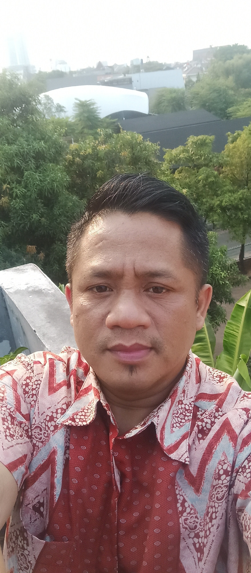 A man in a red patterned collared shirt takes a selfie outdoors. The man has short dark hair and a small goatee. Behind him, lush green trees are visible, and in the distance, several buildings and a large white domed structure are present. The image was taken in West Jakarta, Indonesia.