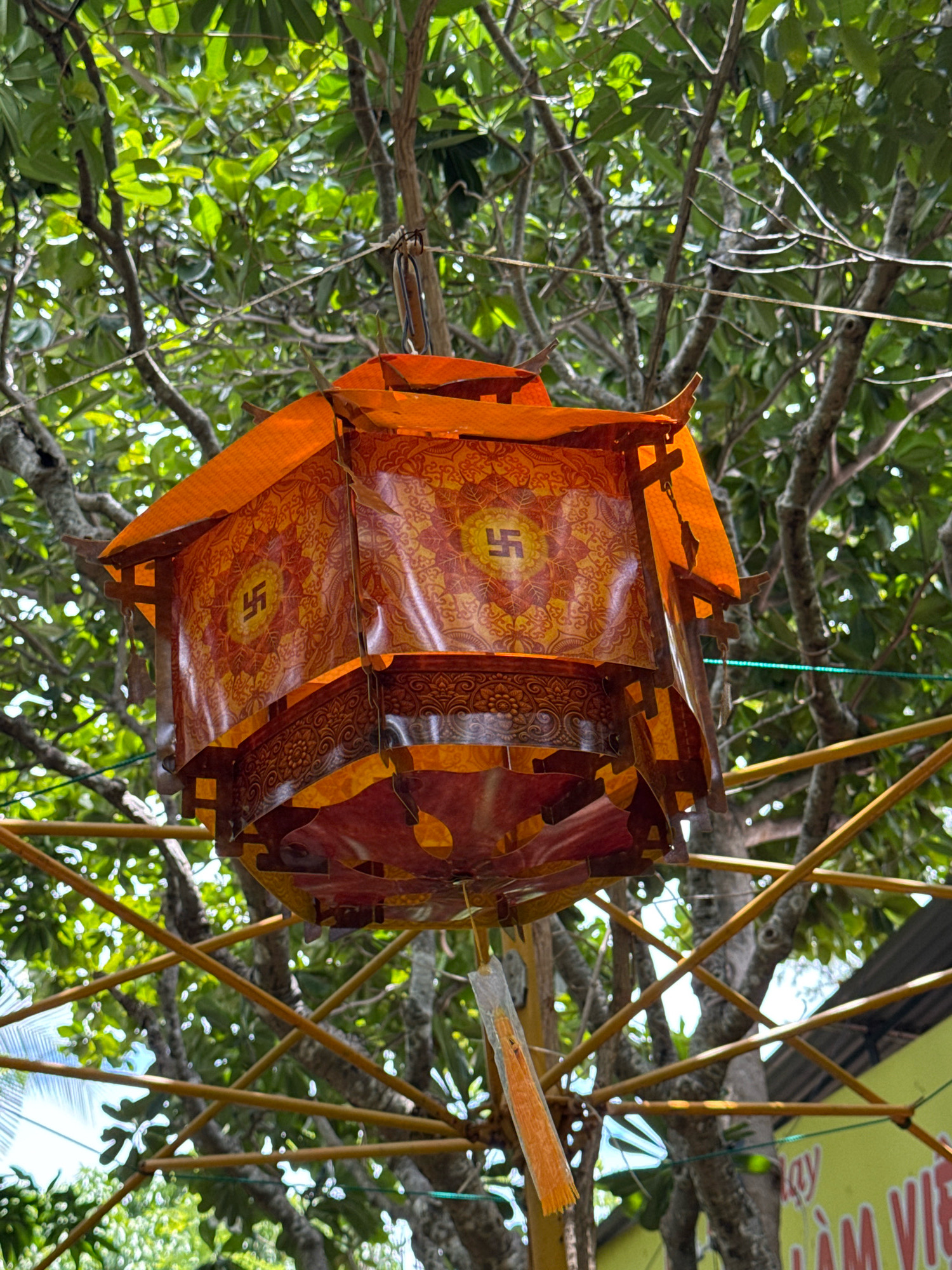 A close-up, eye-level shot captures an ornate, multi-tiered, orange and red paper lantern hanging from a tree branch. The lantern features intricate patterns and a swastika symbol on at least two of its sides, suggesting a cultural or religious significance, likely Buddhist. The background is dominated by the lush green foliage of trees, with sunlight filtering through, creating dappled light. Yellow bamboo-like structures are visible in the lower portion of the frame, crisscrossing behind the lantern. A portion of a yellow sign with Vietnamese text, including the words "LAM VIET", is visible in the far lower right corner, indicating an outdoor location possibly within a market, temple, or cultural site in Nha Trang, Vietnam. There are no people or animals present in the image. The overall impression is one of cultural decoration set against a natural, sunlit backdrop.