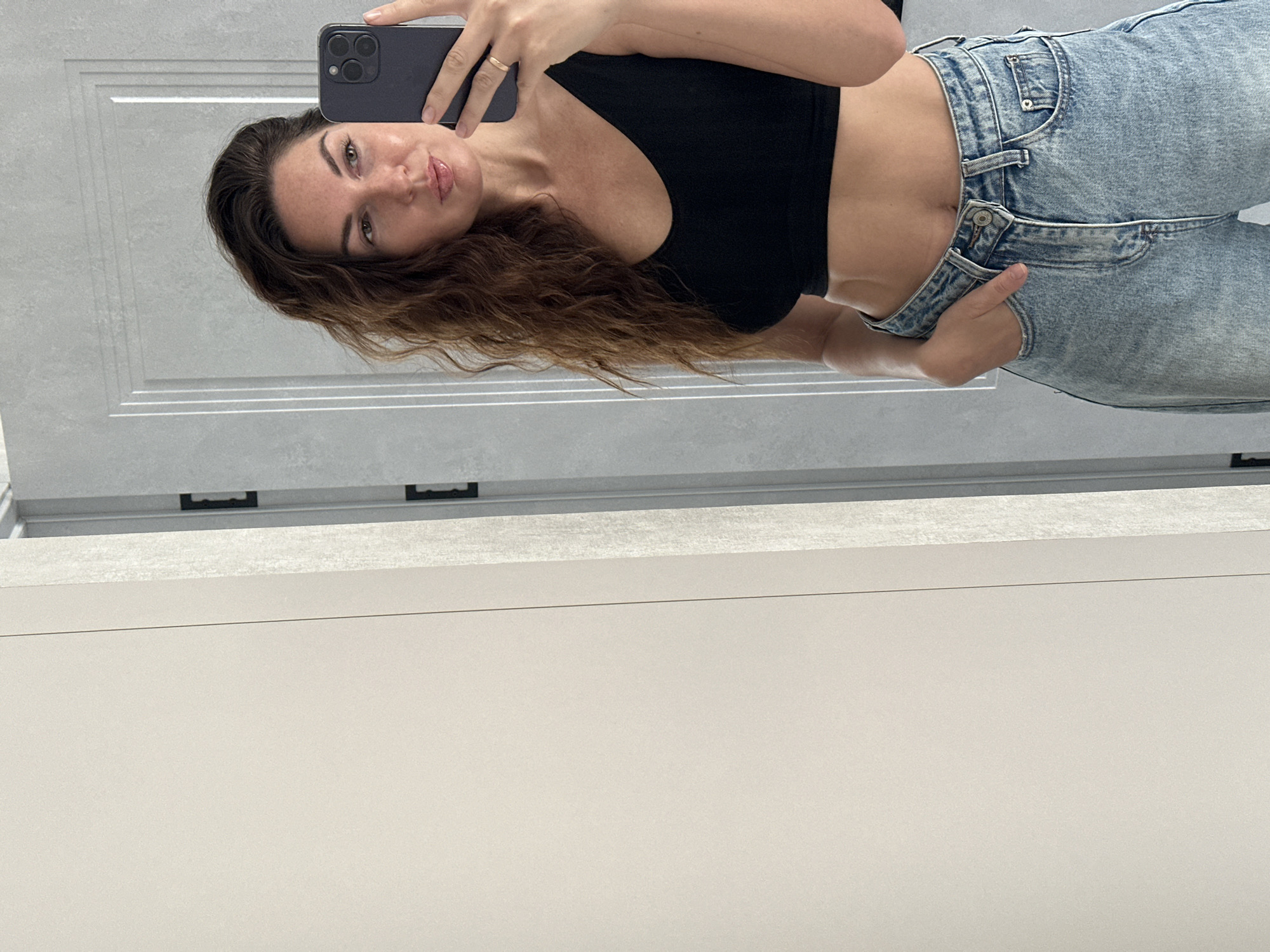 A woman is taking a mirror selfie. She is wearing a black crop top and light blue denim jeans. Her hand is in her pocket and she is looking at her phone. She has long, wavy brown hair and is wearing a ring on her right hand. The background appears to be a doorway with decorative paneling. The image was taken in a bathroom, likely in Chaikovsky, Russia.